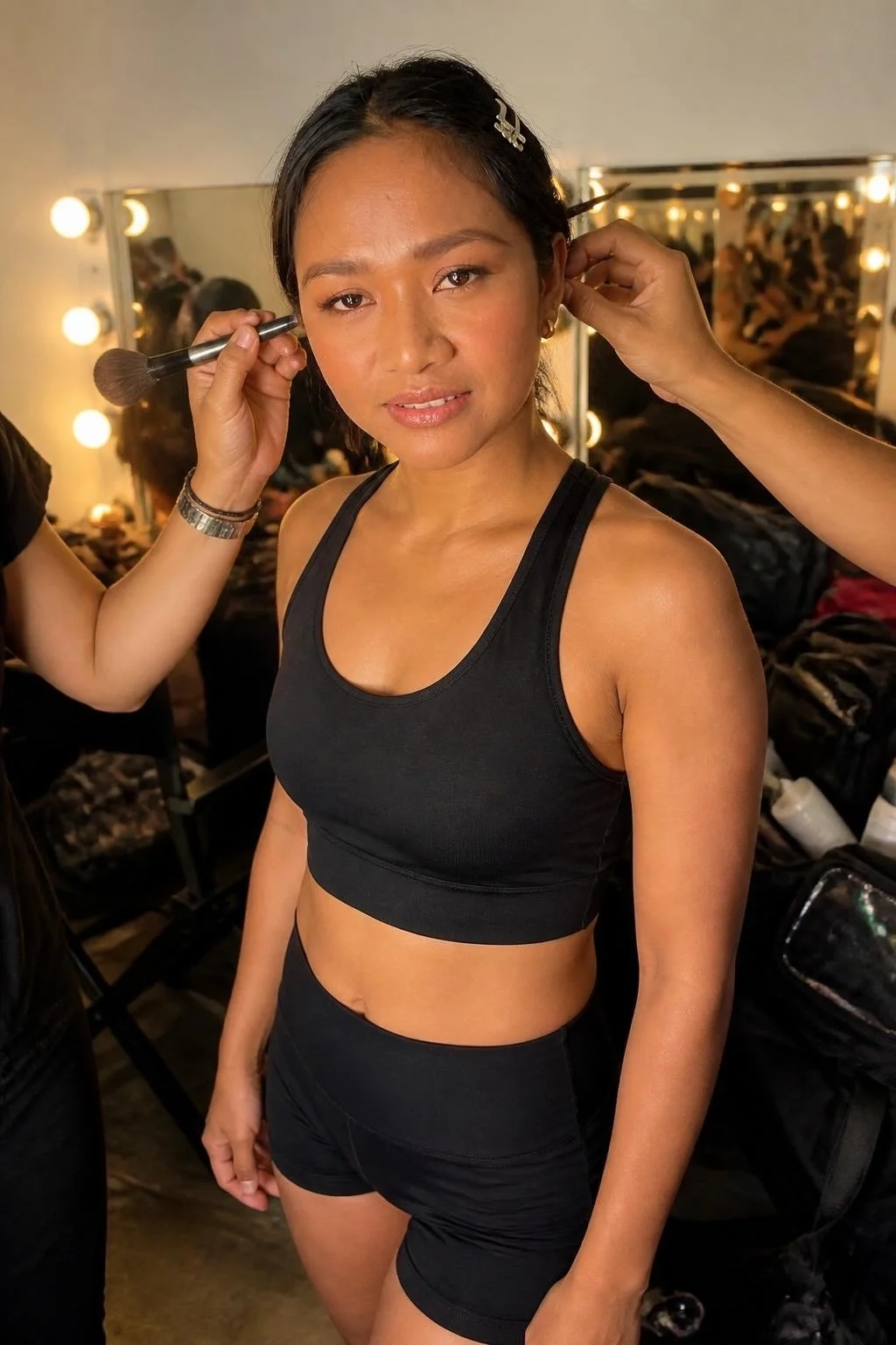A woman with medium skin tone and black hair having makeup applied by a makeup artist in a dressing room with bright vanity lights.
