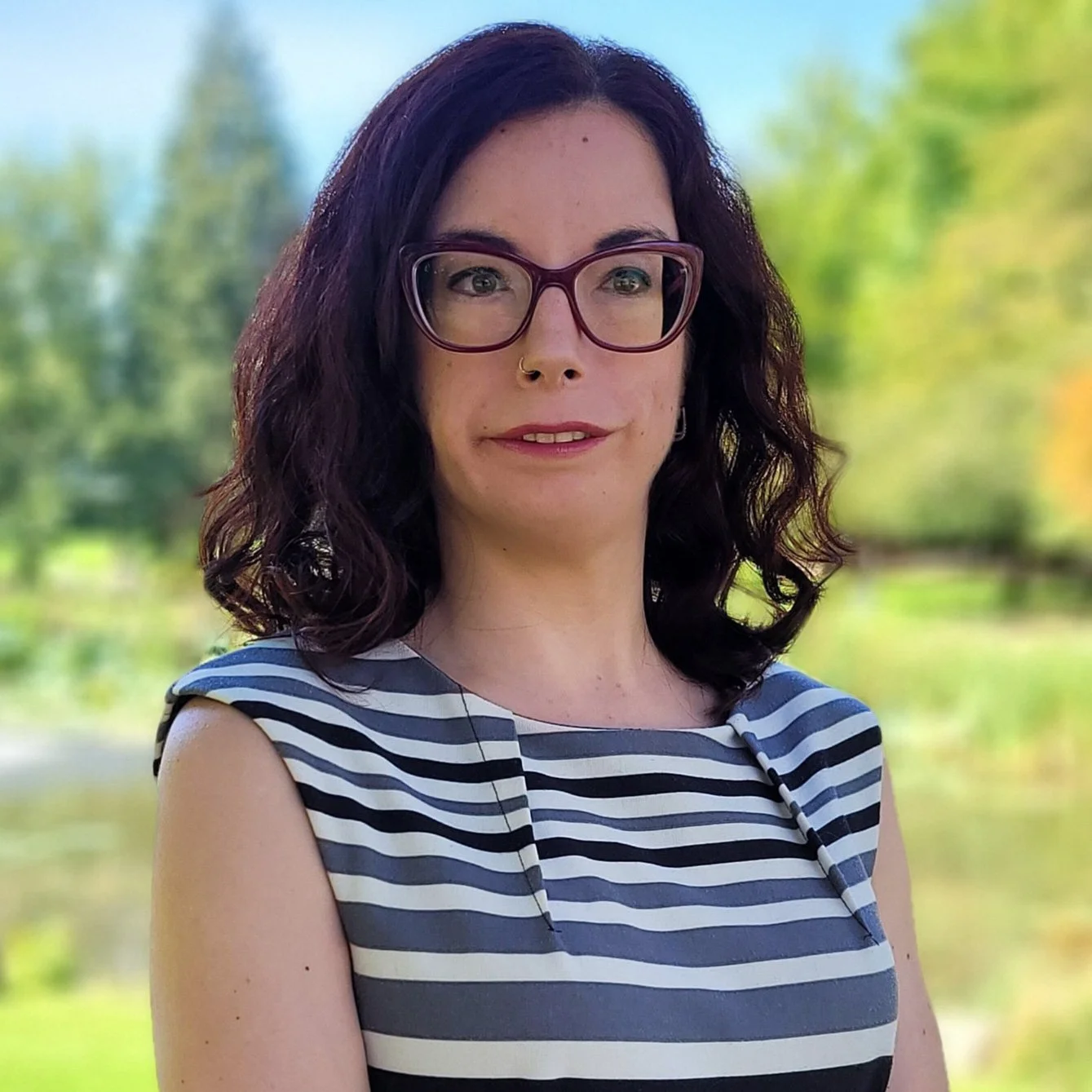 A woman with dark, shoulder-length curly hair, wearing glasses, a nose ring, and a sleeveless striped dress, standing outdoors with a blurred green and yellow background.