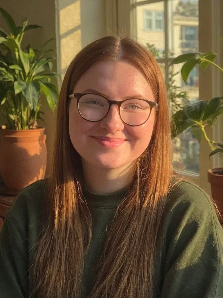 A young woman with long brown hair and glasses smiling indoors in front of large windows with sunlight and houseplants.
