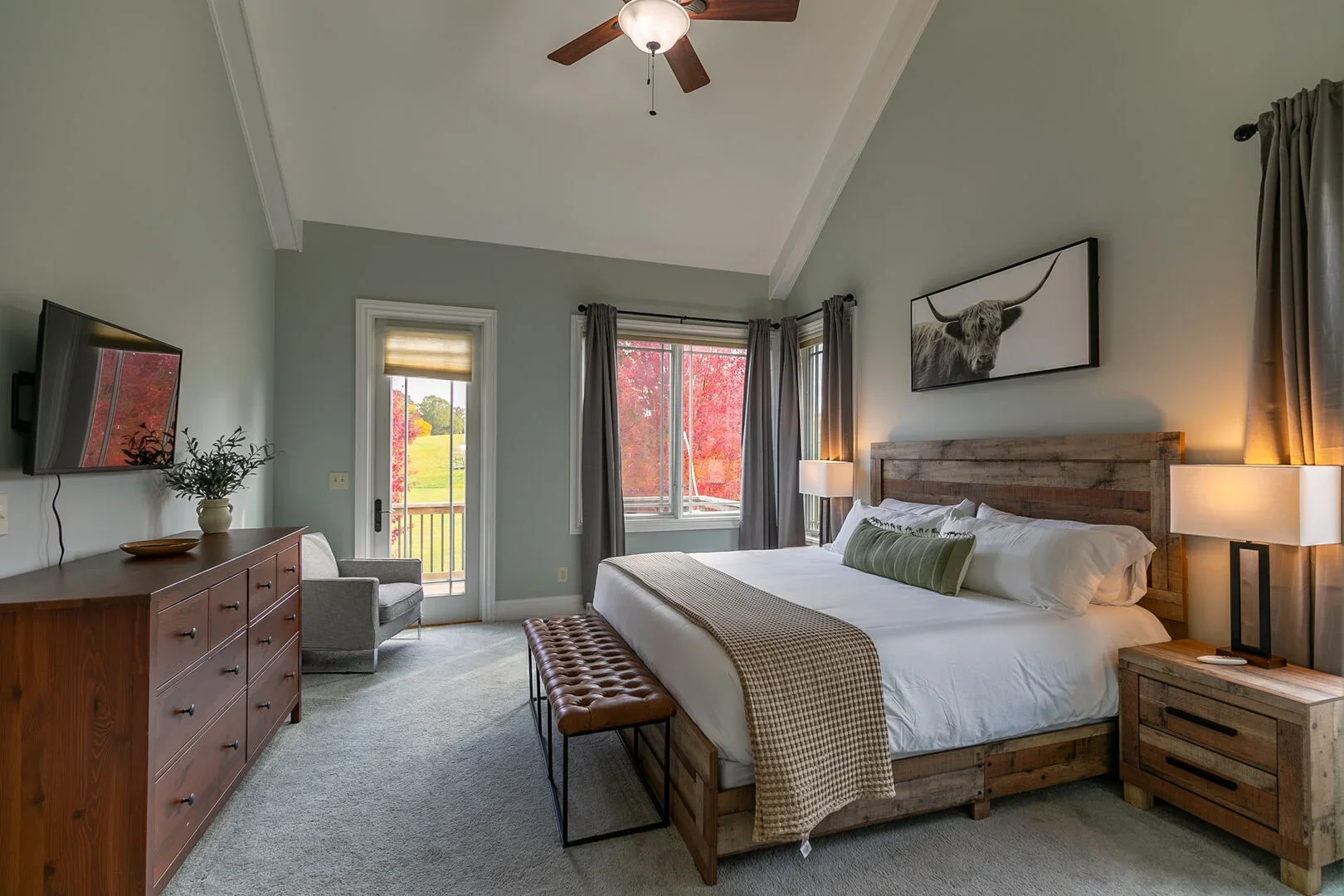 A spacious bedroom with a large bed, wooden headboard, and nightstands, illuminated by table lamps, with large windows showing a vibrant outdoor landscape with red foliage.