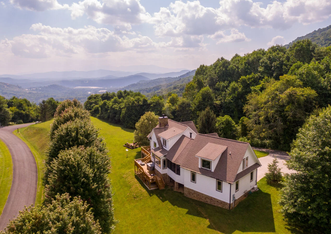 A white two-story house with multiple gable roofs surrounded by green trees and rolling hills under a partly cloudy sky.