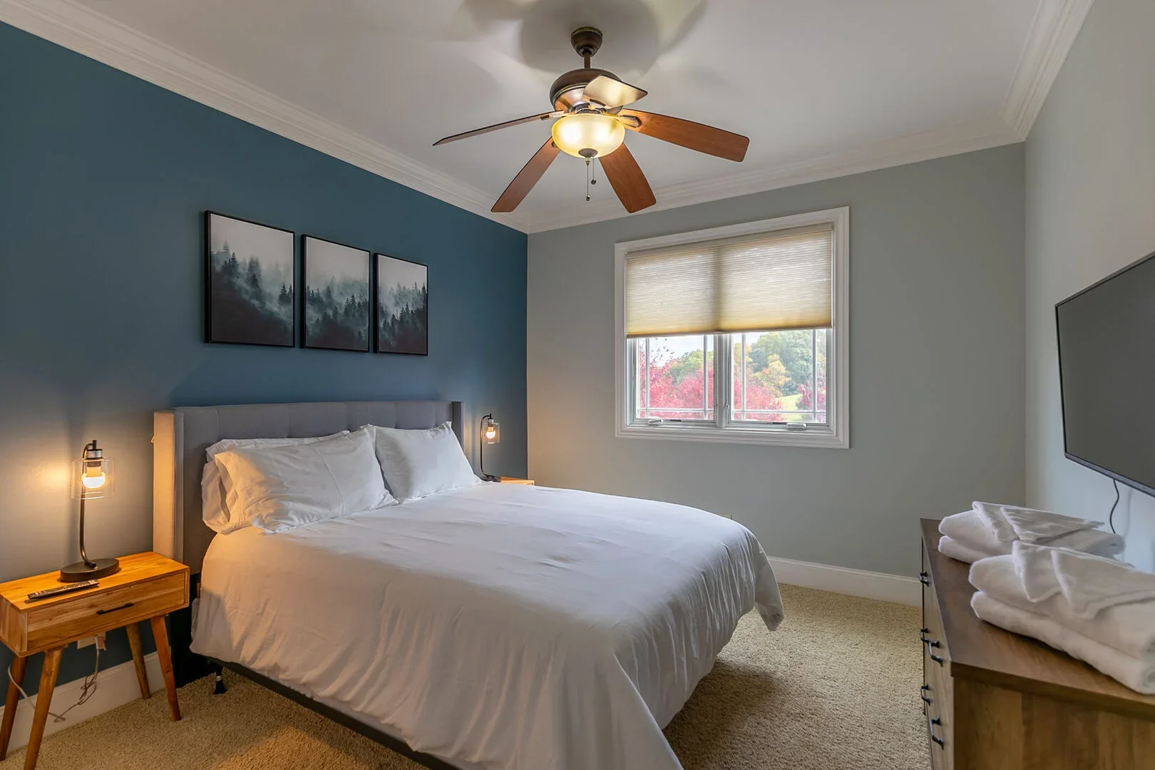 A bedroom with a bed, wooden nightstand with a lamp, window with a blind, and a wall-mounted TV.