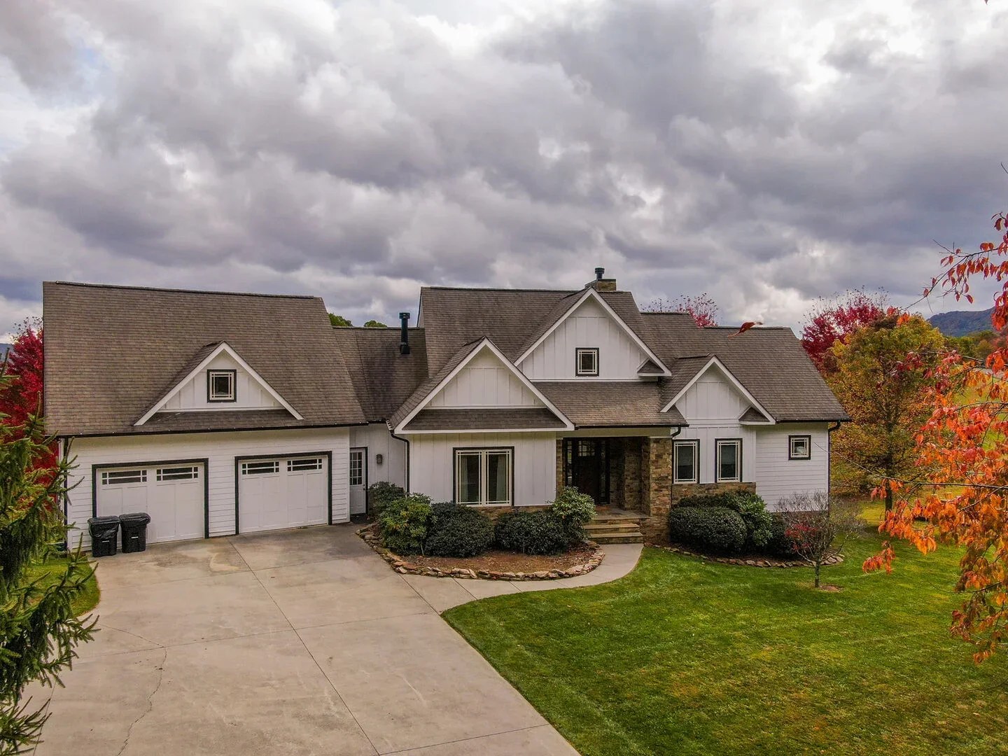 A large white house with a gray roof, attached two-car garage, front steps, and landscaped front yard with bushes and trees with orange and red leaves, under a cloudy sky.