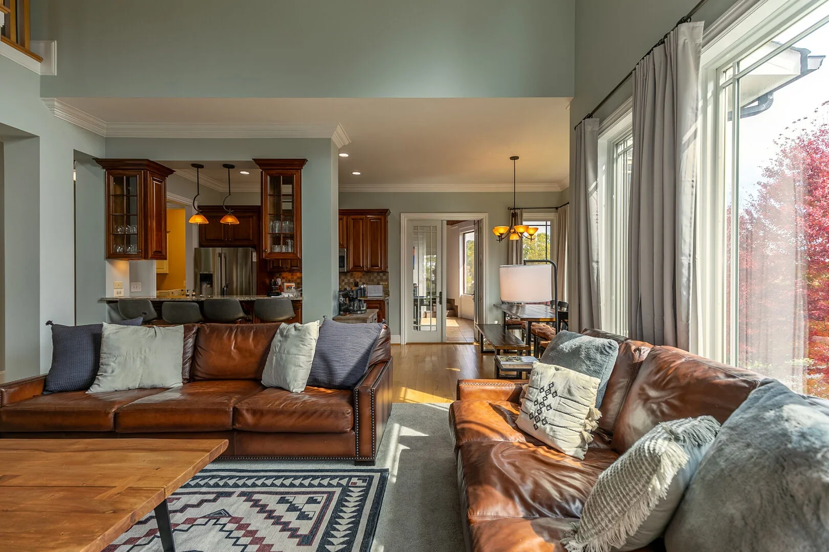 Living room with large windows, leather sofas, decorative pillows, a rug, and an open kitchen with bar stools in a home interior.