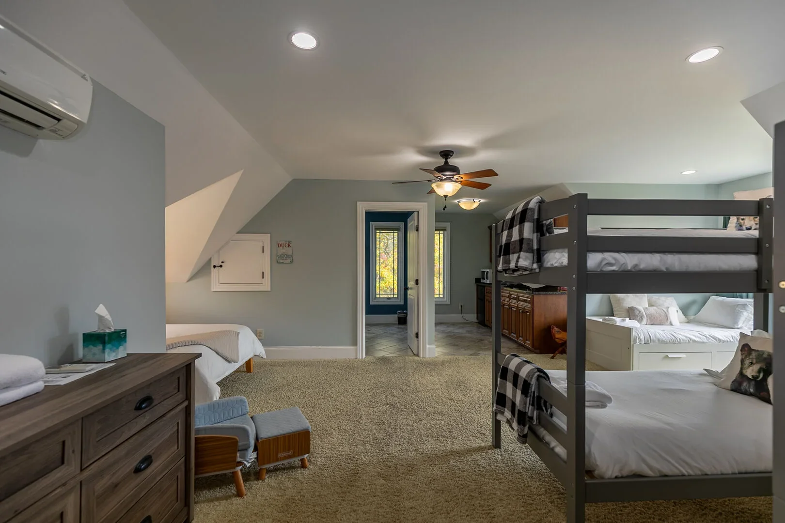 Interior of bedroom with bunk beds, single bed with dog pillow, dresser with tissues, small chairs, ceiling fan, air conditioning, and a door leading to a room with a window showing trees outside.