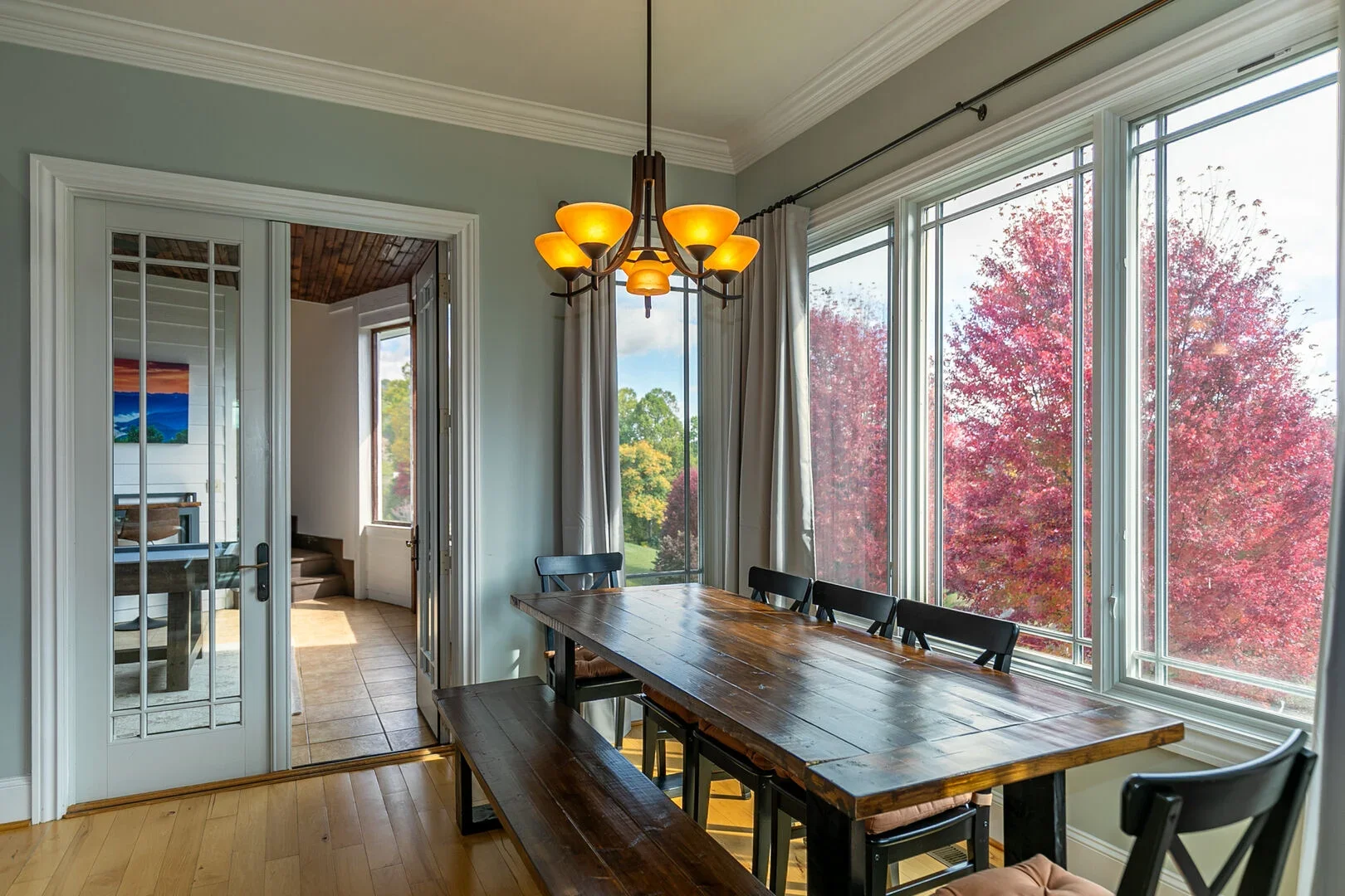 Dining area with a wooden table, black chairs, and large windows showing red and green autumn trees outside, with a chandelier hanging above.