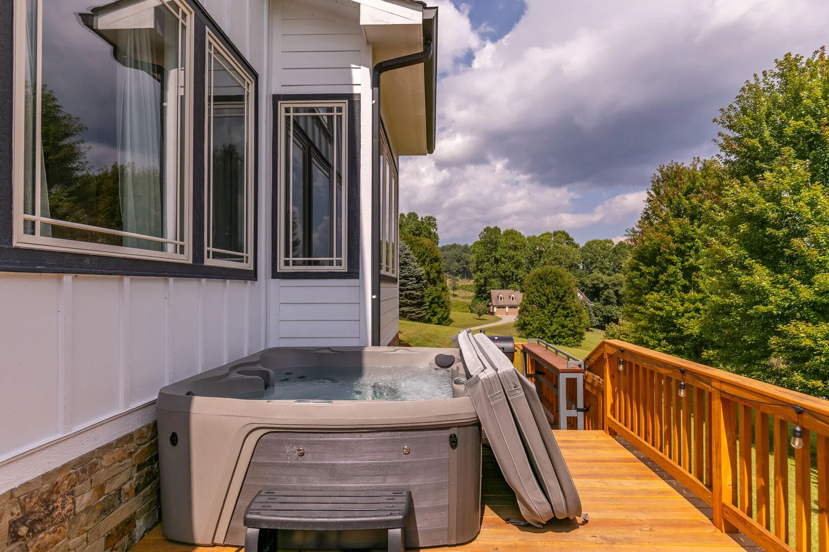 Hot tub on a wooden deck outside a house, with trees and a partly cloudy sky in the background.