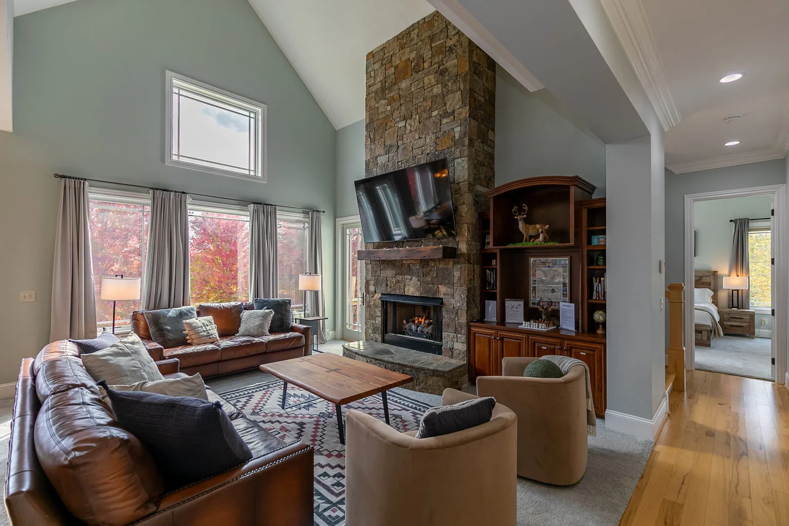 Living room with a stone fireplace, wall-mounted TV, leather sofas, armchairs, a wooden coffee table, and large windows showing autumn trees.