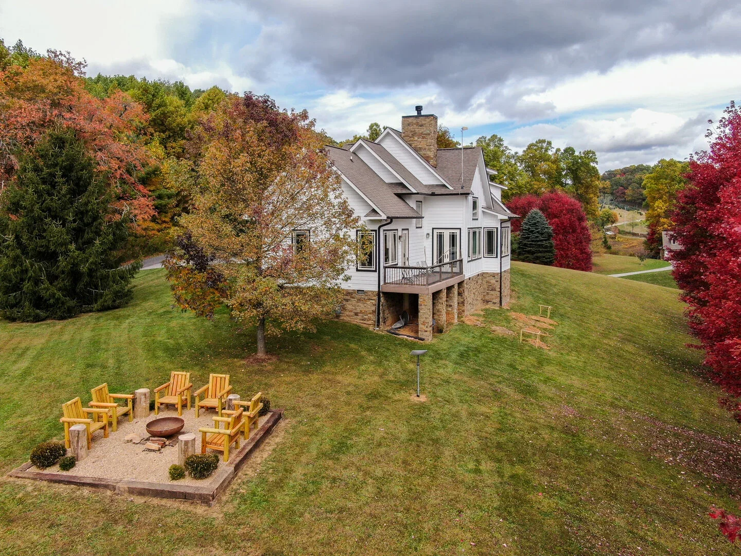 A large white house with multiple gabled roofs and a stone foundation is situated on a grassy hill with autumn-colored trees around it. There is a small wooden deck at the back, and a fire pit with wooden chairs around it on the lawn.