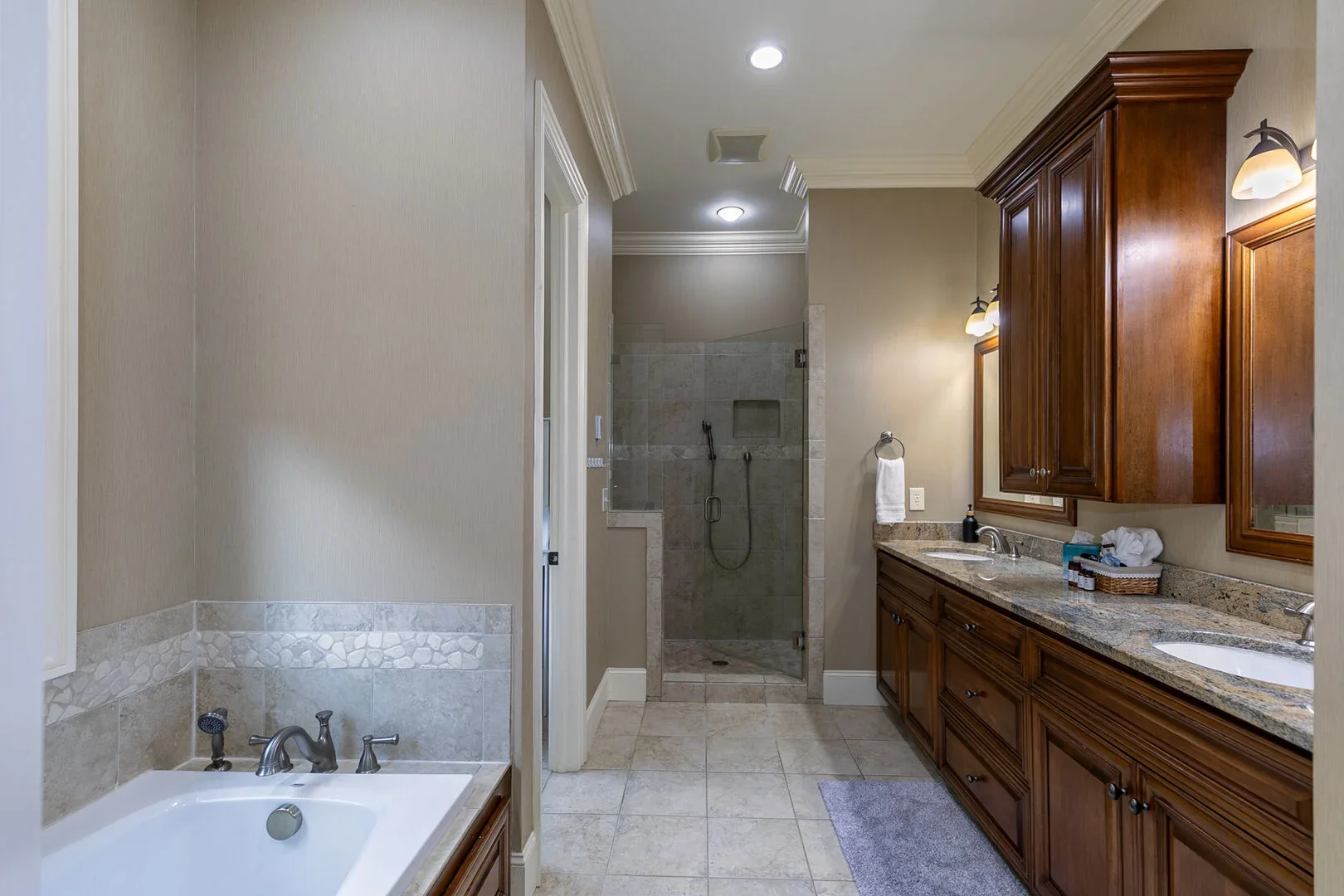 Bathroom with double vanity, large mirror, granite countertops, wooden cabinetry, shower with glass door, bathtub, and beige tile flooring.