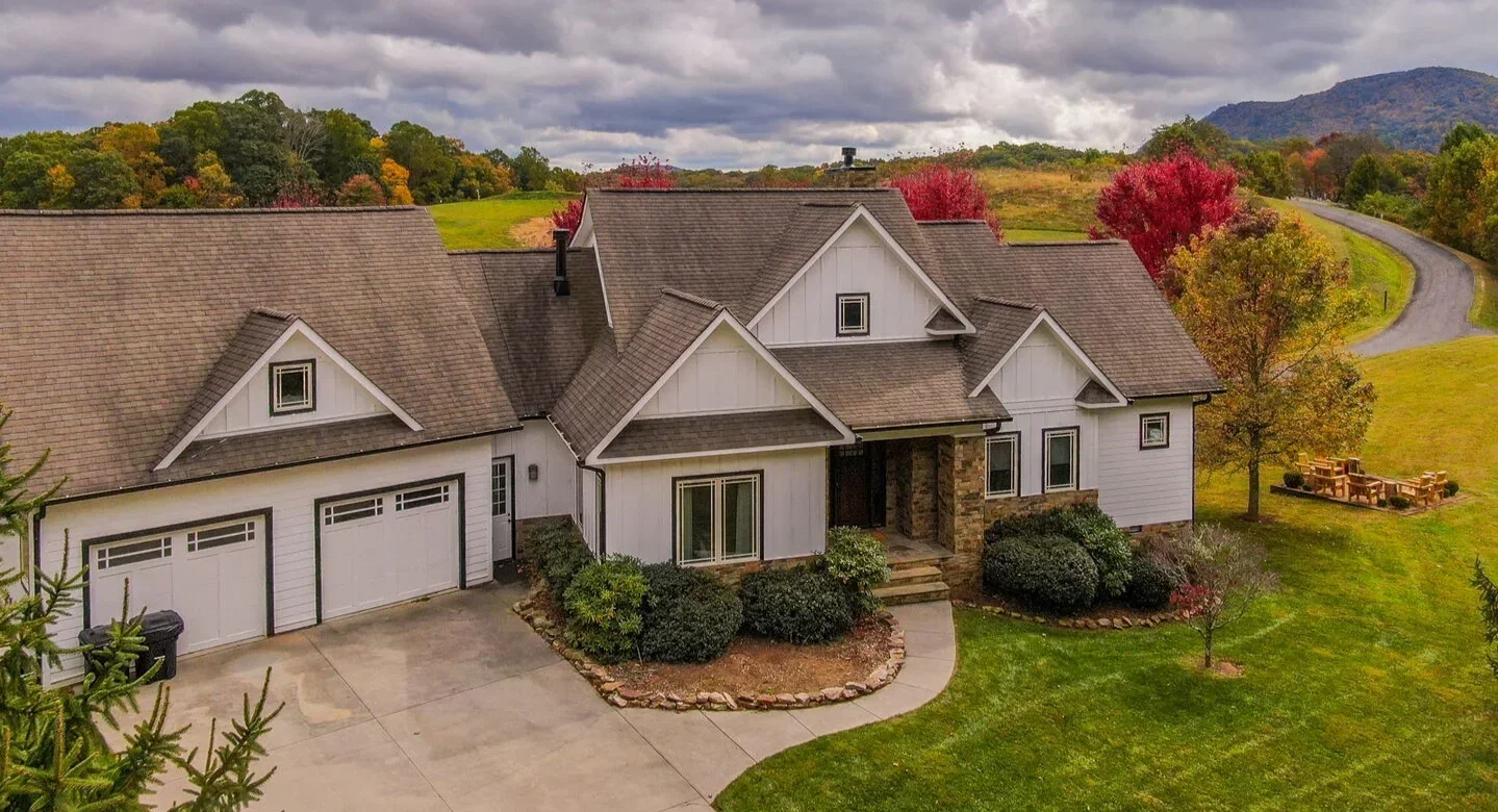 A large two-story house with a gray shingle roof and white exterior, surrounded by a green lawn with trees displaying fall colors, and a winding driveway leading to a grazing pasture.