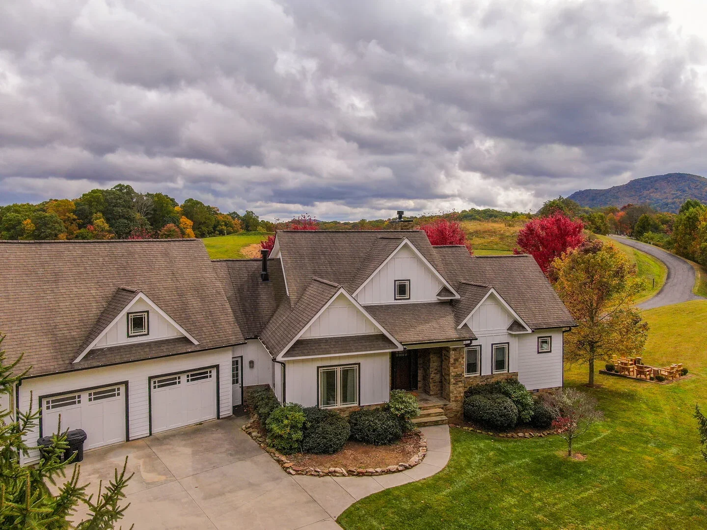 A large white house with a brown shingle roof, surrounded by a spacious lawn and colorful autumn trees, with a winding driveway and mountains in the background under a cloudy sky.