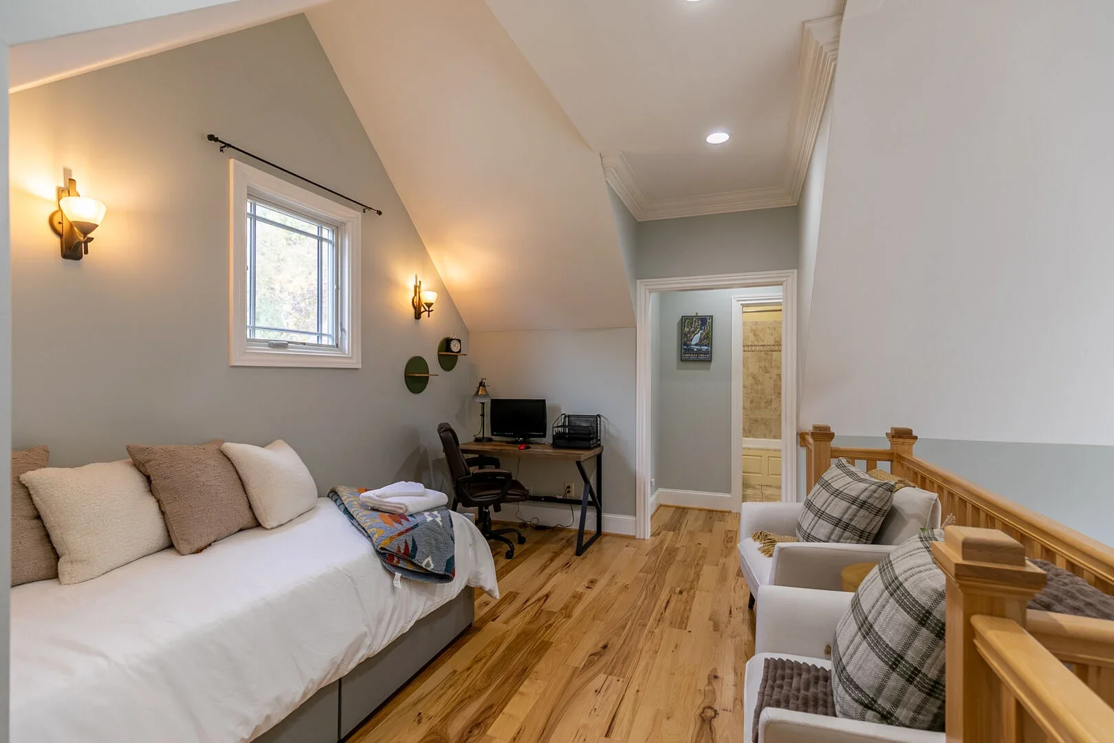 Living space with a window, wall-mounted lights, a couch with pillows and a blanket, a small desk with a computer and chair, and two armchairs with plaid pillows. There's an open door leading to another room.