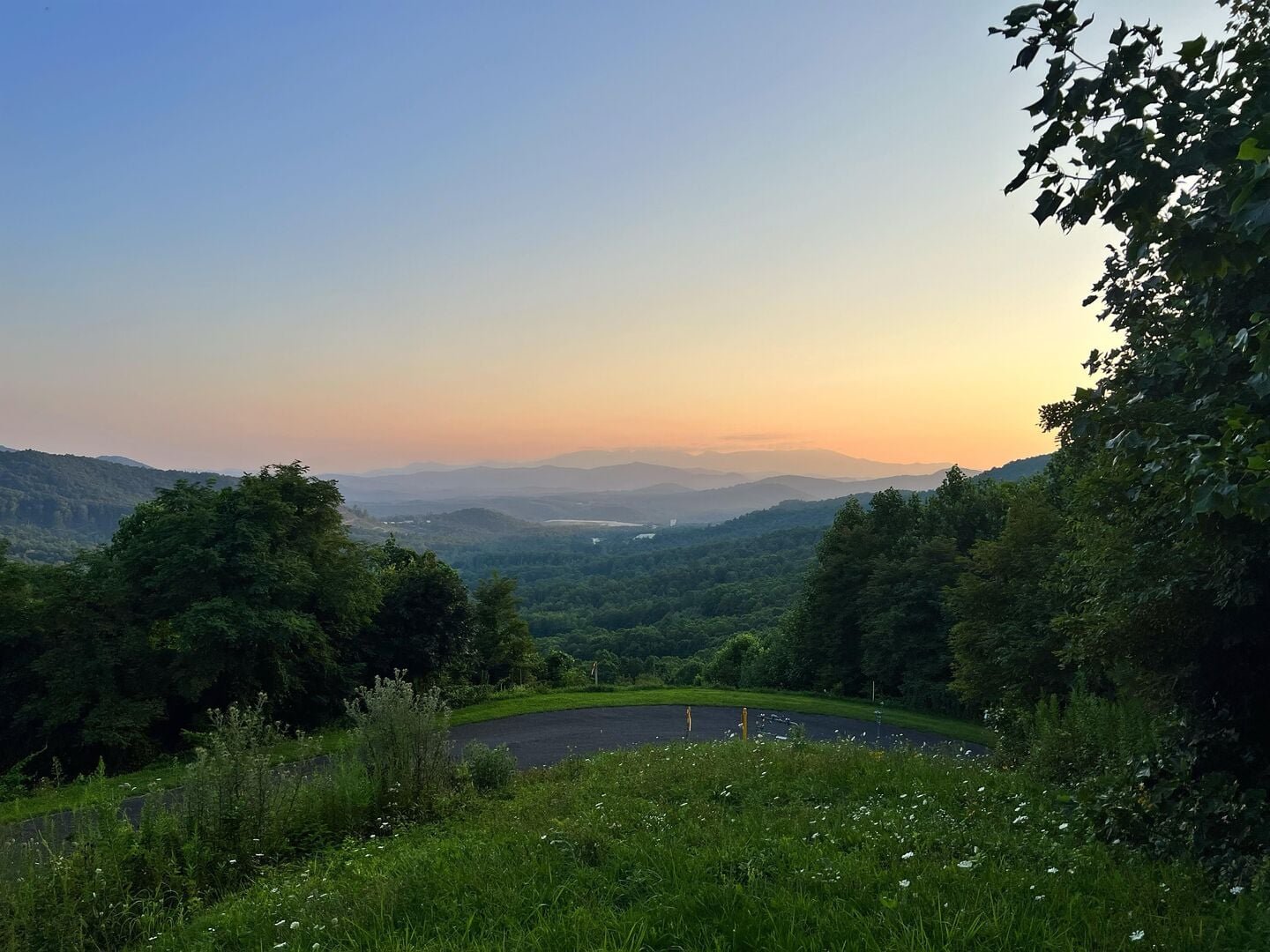 A scenic view of rolling green hills and distant mountains at sunset, with a curved road in the foreground and trees on the sides.