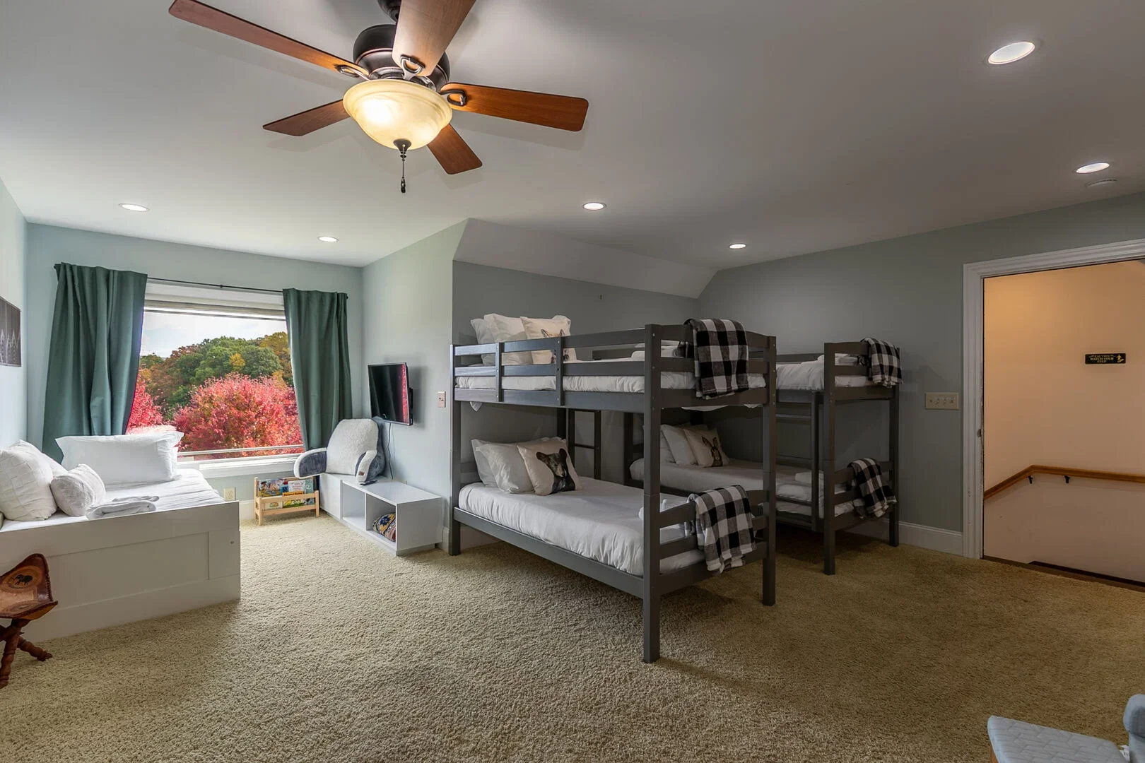 A bedroom with bunk beds, a couch near a window with green curtains, a small white bookshelf, a wall-mounted TV, and a ceiling fan. Outside the window are trees with pink, red, and green foliage.