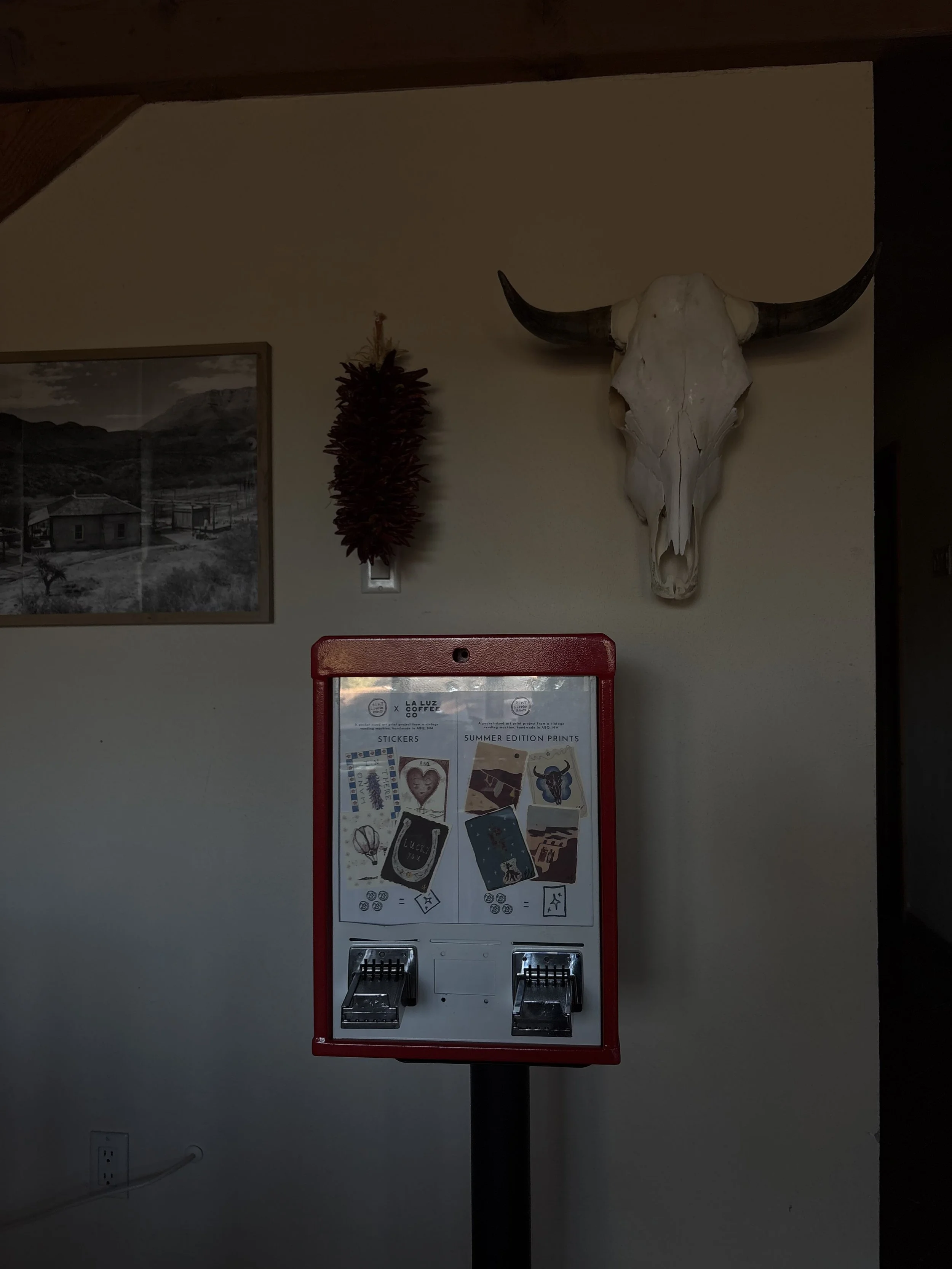 Indoor scene with a decorative cow skull mounted on the wall, a framed black and white landscape photograph, and a red vending machine or display stand with stickers and printings.