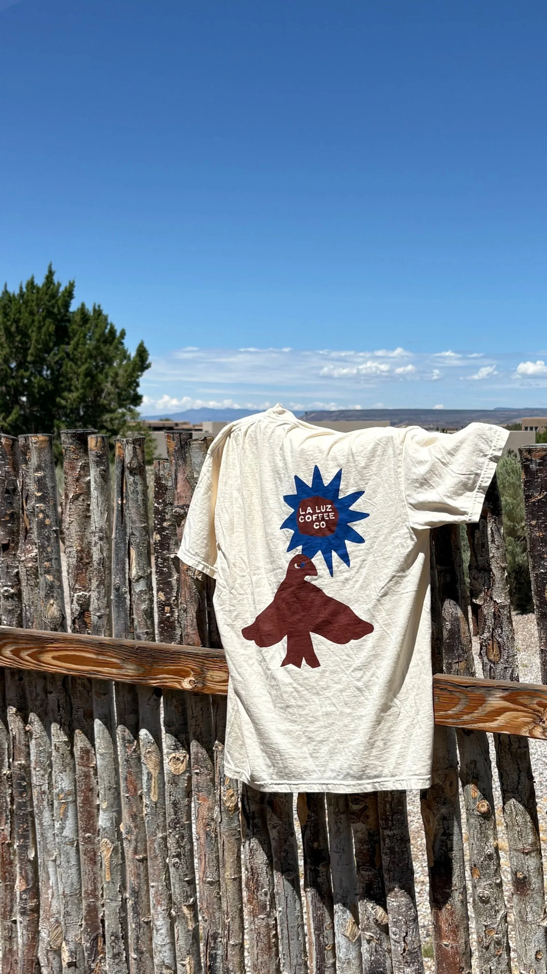 A white t-shirt with a graphic design hanging on a rustic wooden fence outdoors under a blue sky with some clouds, and a tree to the left.