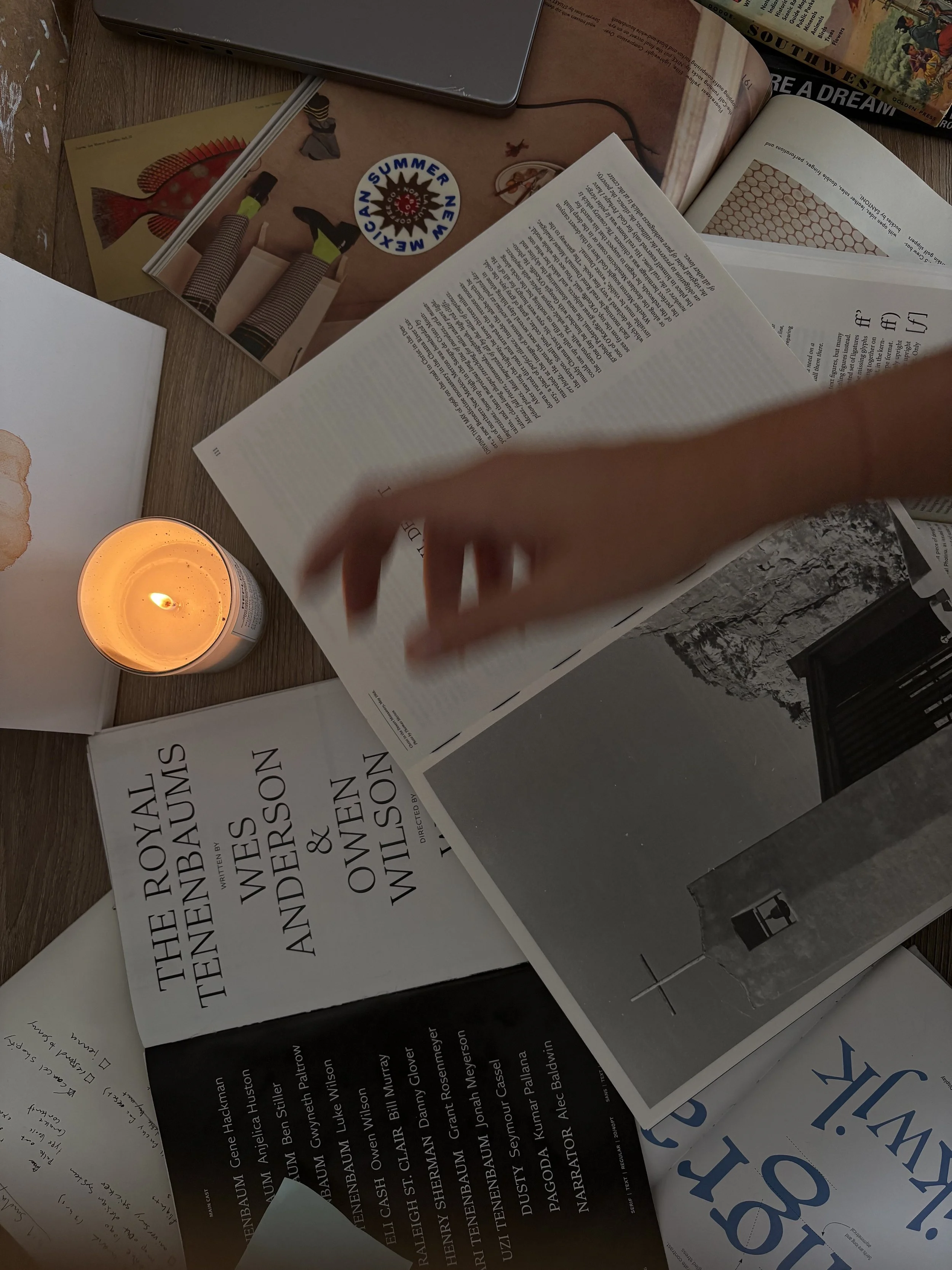 A cluttered desk with open books, papers, a lit candle, and a laptop.