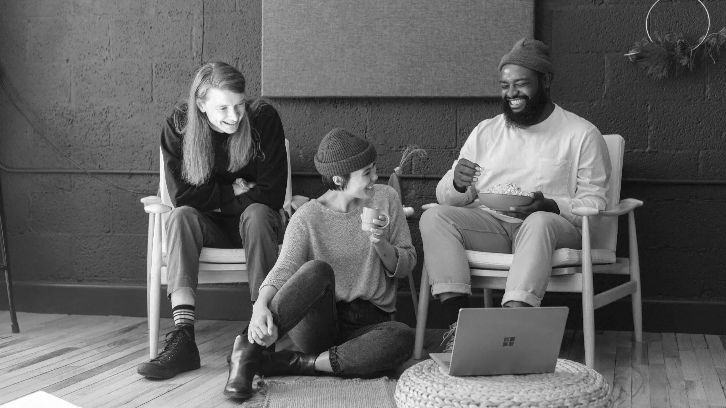 Four friends sitting and chatting, one person holding a bowl, another with a mug, and a laptop on a woven table, in a cozy indoor setting.