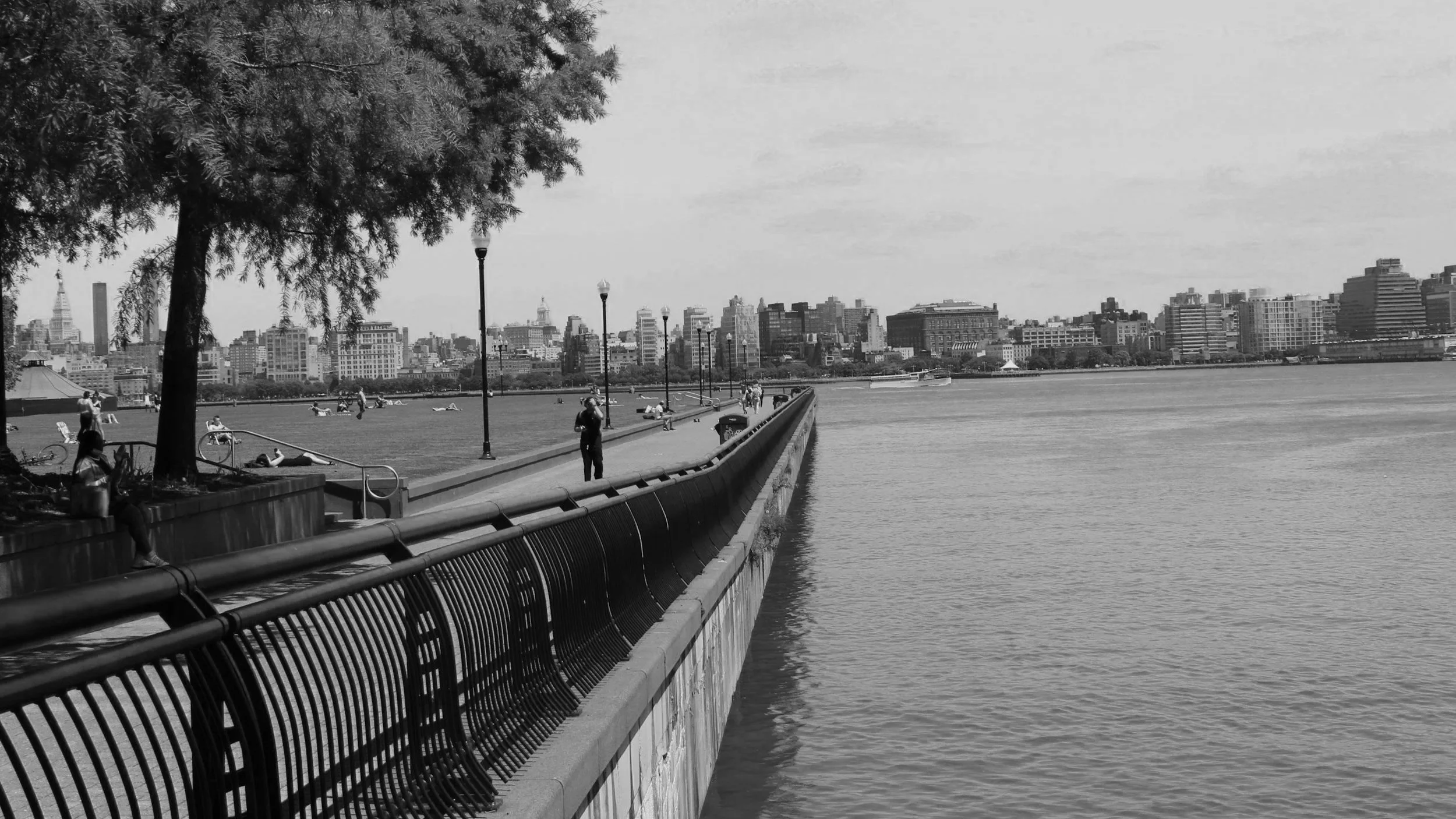 A black and white photo of a waterfront walkway with people and city buildings in the background.