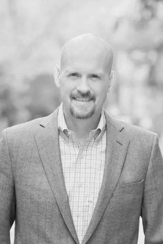 A black and white photo of a bald man with a mustache and beard, wearing a checked shirt and blazer, smiling outdoors.