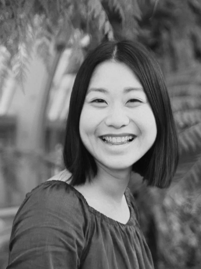Black and white portrait of a smiling Asian woman with shoulder-length hair, wearing a gathered neckline top, in front of greenery.