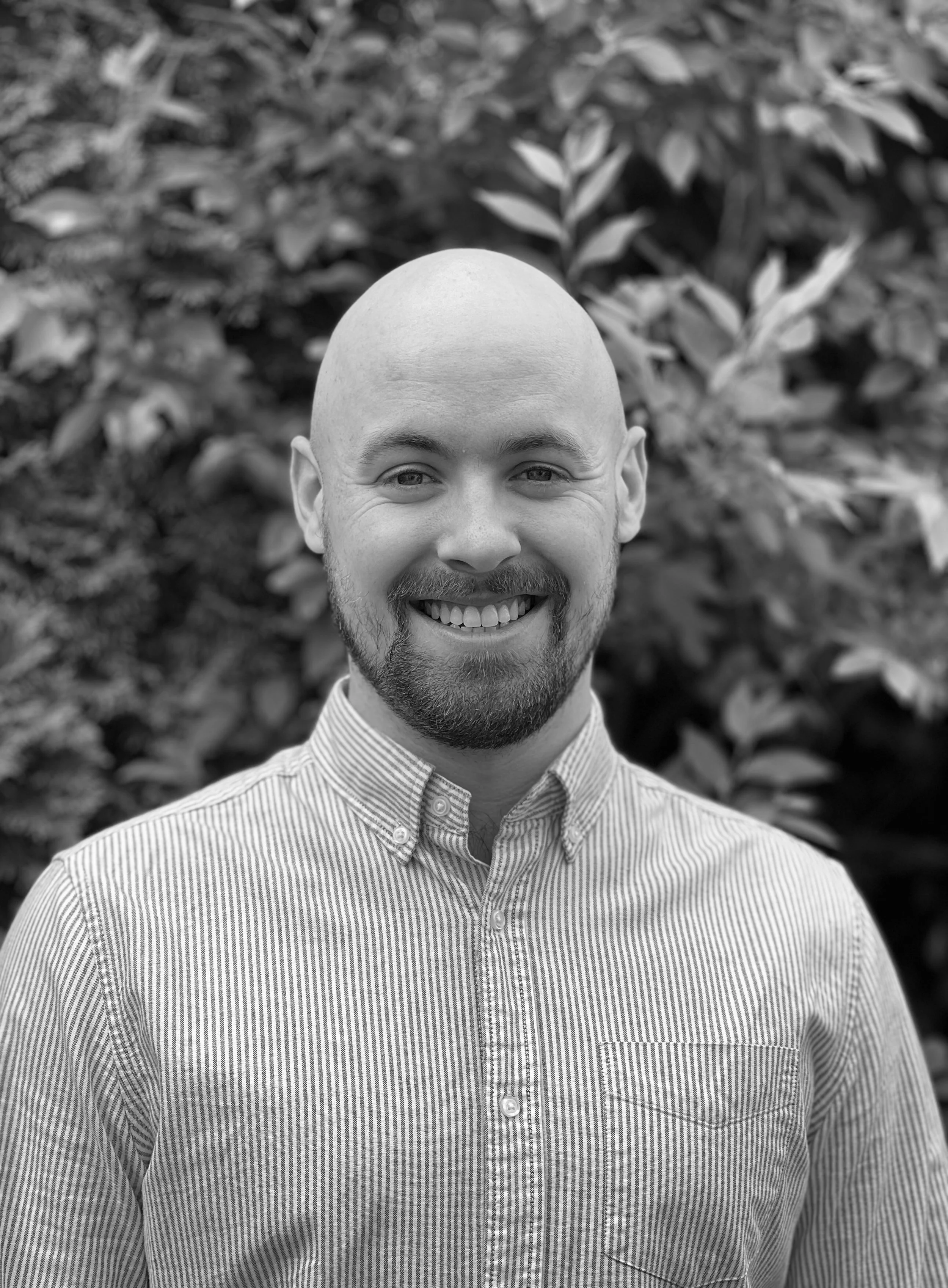 A smiling man with a bald head and beard wearing a striped button-up shirt standing outdoors in front of foliage.