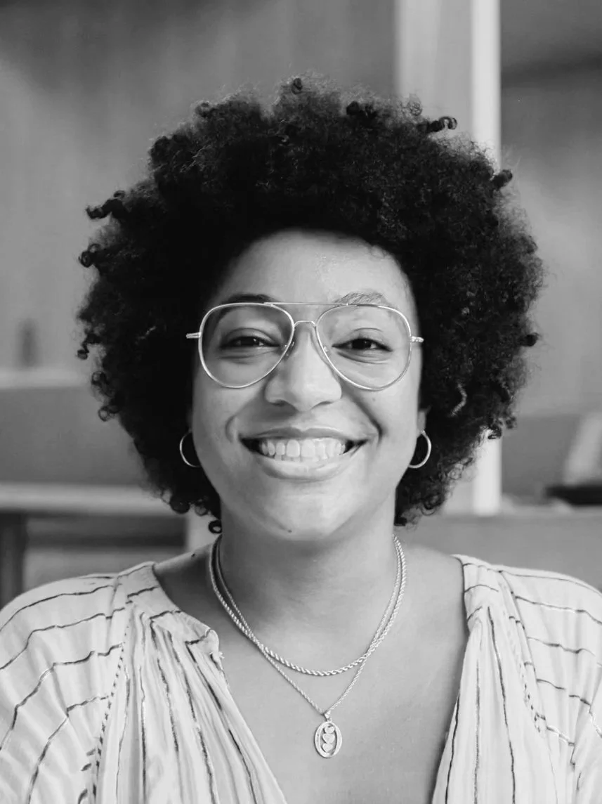 A woman with curly hair, wearing glasses, smiling, dressed in a striped top, with hoop earrings and layered necklaces, in an indoor setting.