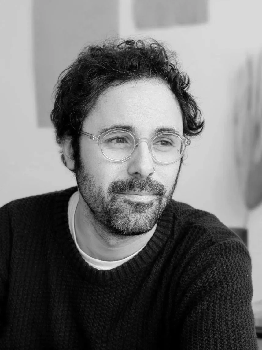 A black and white photo of a man with glasses, a beard, and curly hair, wearing a dark sweater, looking to the side with a slight smile.