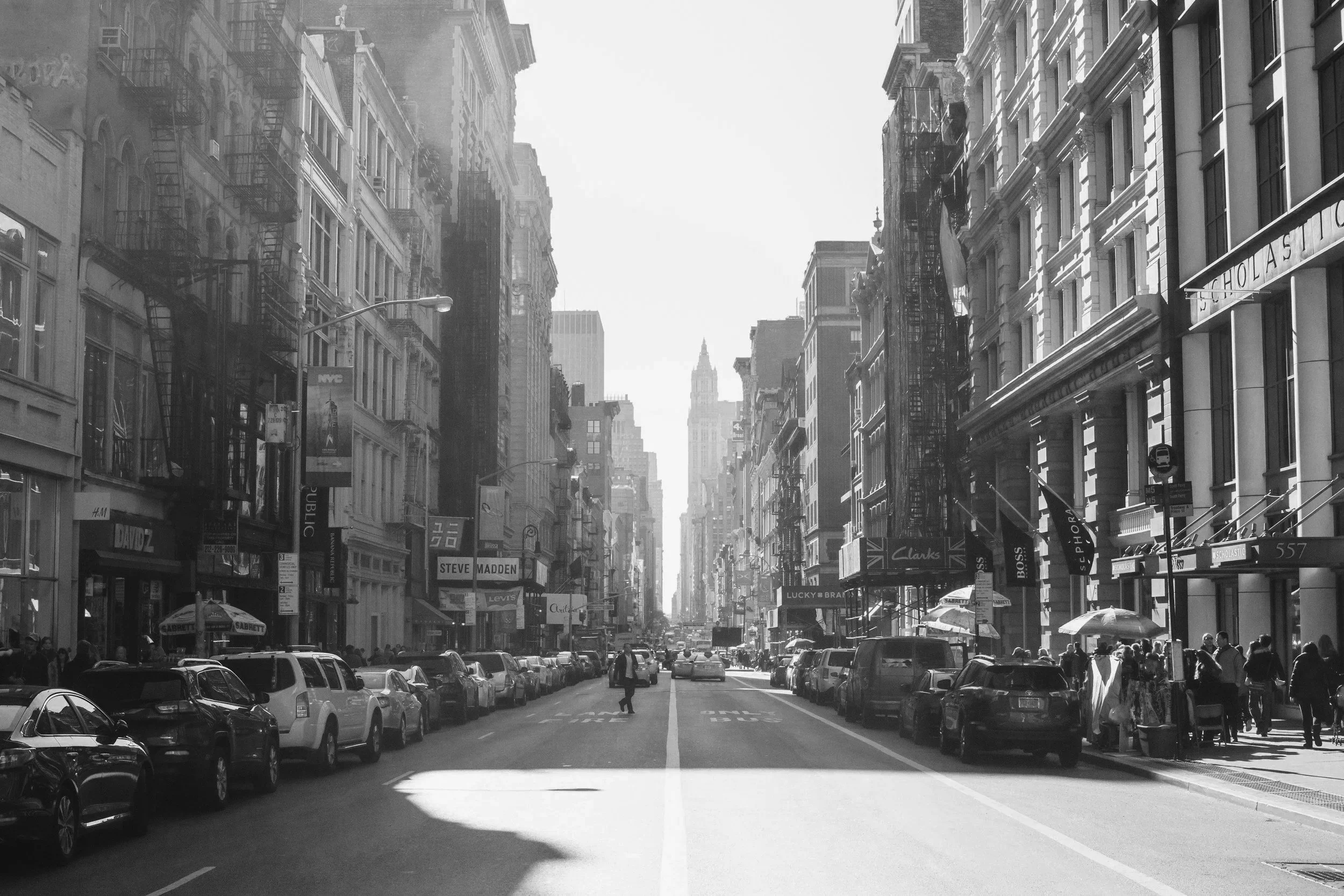A city street view with cars parked on both sides, storefronts, pedestrians, and tall buildings with some fire escapes, with a tower visible in the distance.