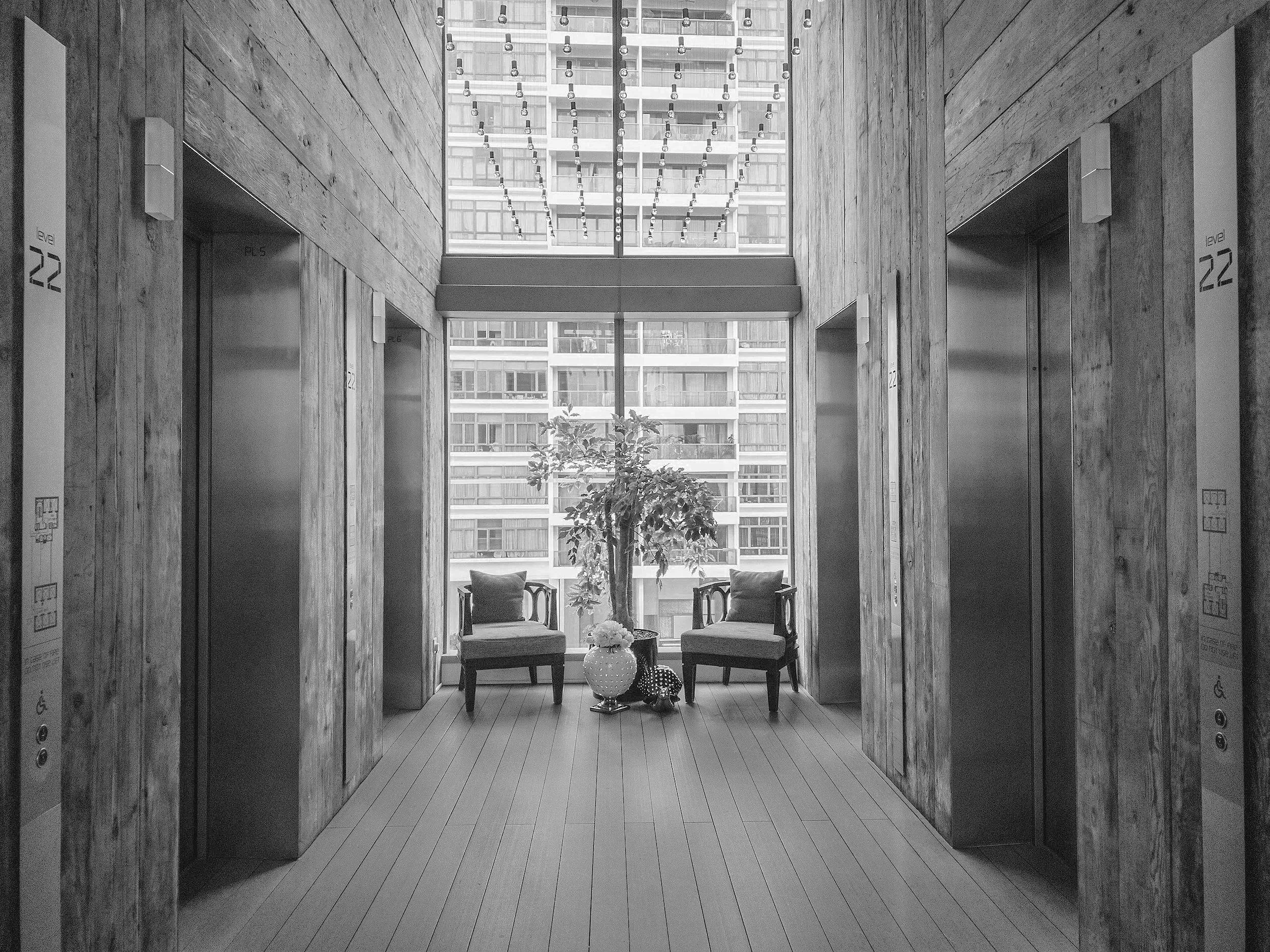 Indoor elevator lobby with wood-paneled walls and two elevator doors on each side. Two chairs with cushions are set in front of a large window showing an apartment building exterior. A potted plant and decorative item sit on the floor between the chairs.