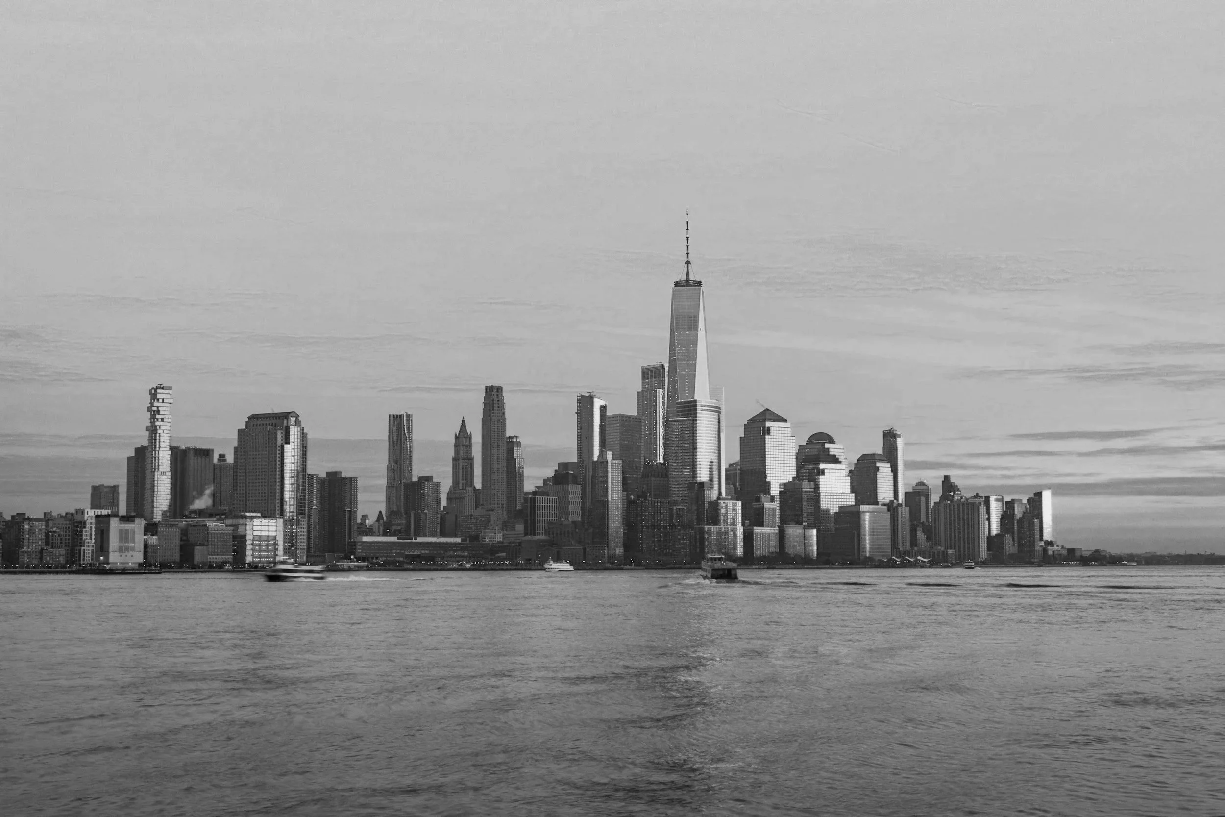 A grayscale image of the Manhattan skyline viewed across a body of water with boats.