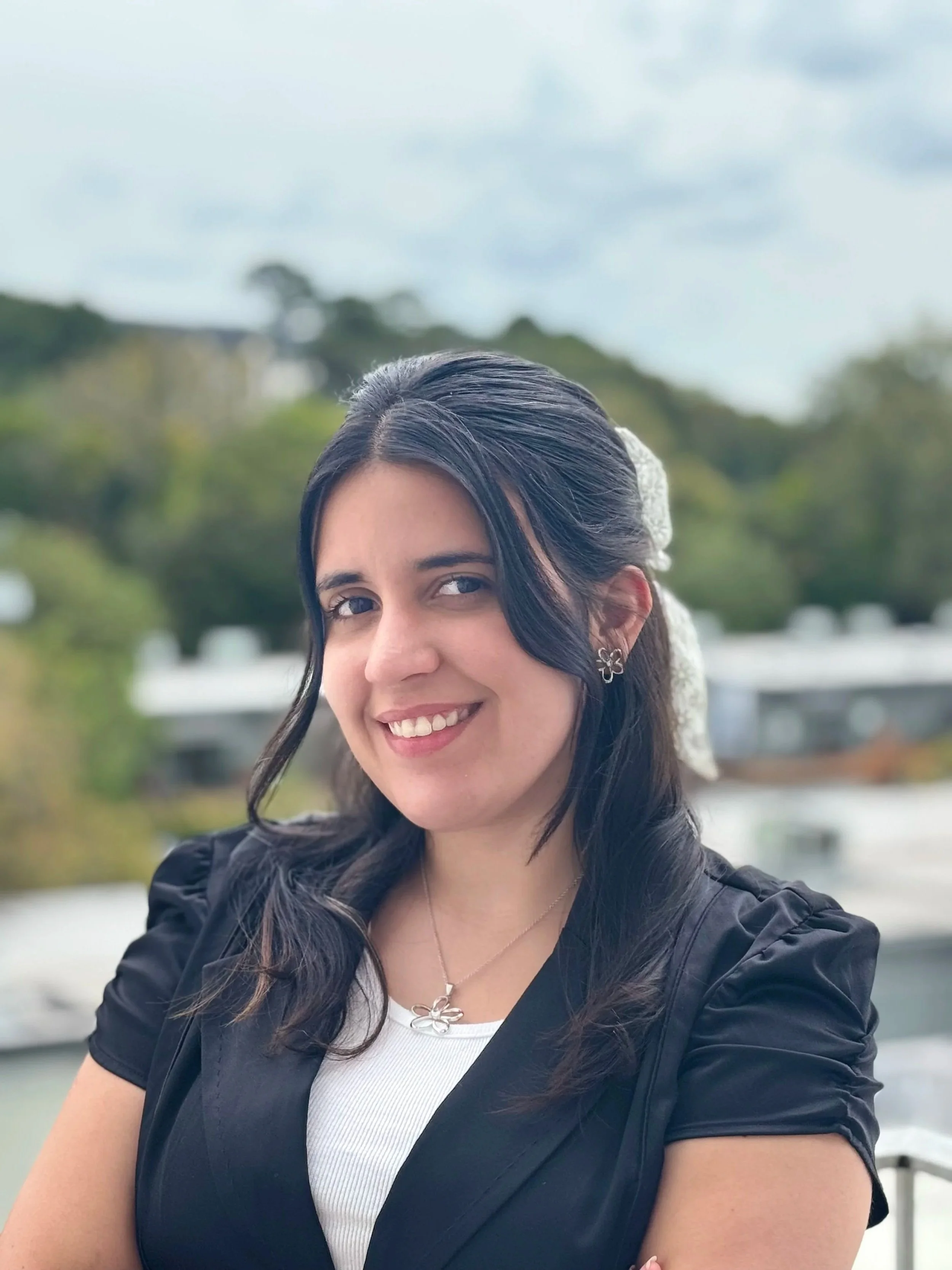 A young woman with long dark hair, smiling, wearing earrings and a necklace, with outdoor scenery in the background.