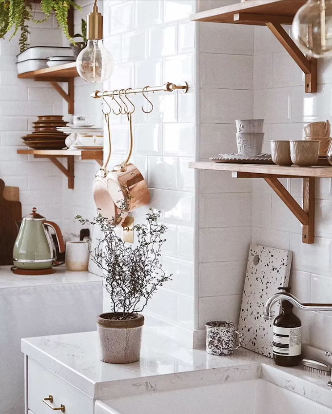 A kitchen scene with wooden shelves holding dishes and bowls, a copper pot hanging, a potted plant on a white counter, and a kettle. The background features white subway tiles, with hanging light fixtures and a minimalistic, modern aesthetic.