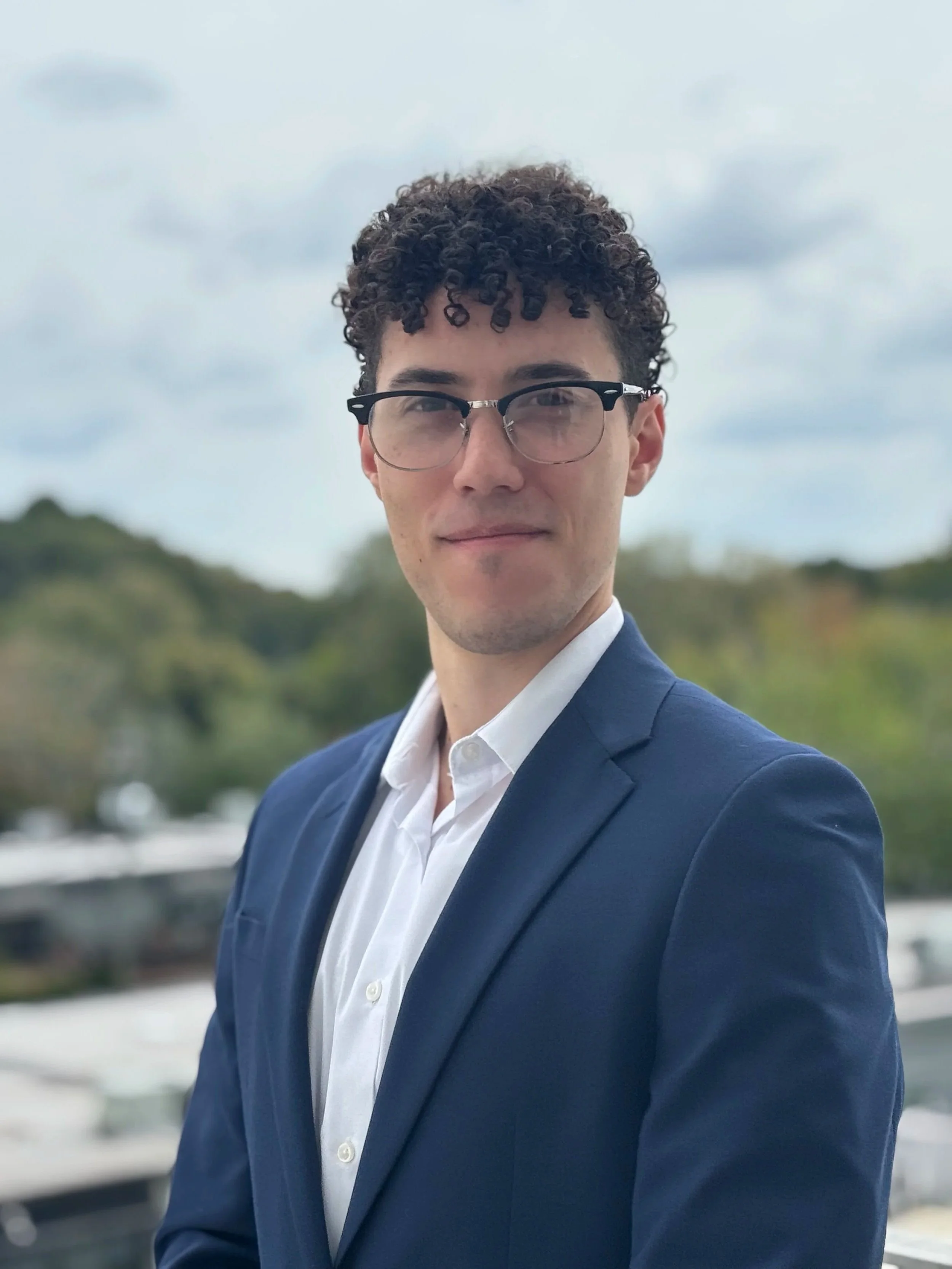 A young man with curly hair and glasses wearing a blue suit and white shirt, outdoors on a cloudy day.