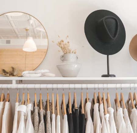 Retail display with clothing on hangers, a round mirror, a white vase with dried flowers, and a black hat on a stand.