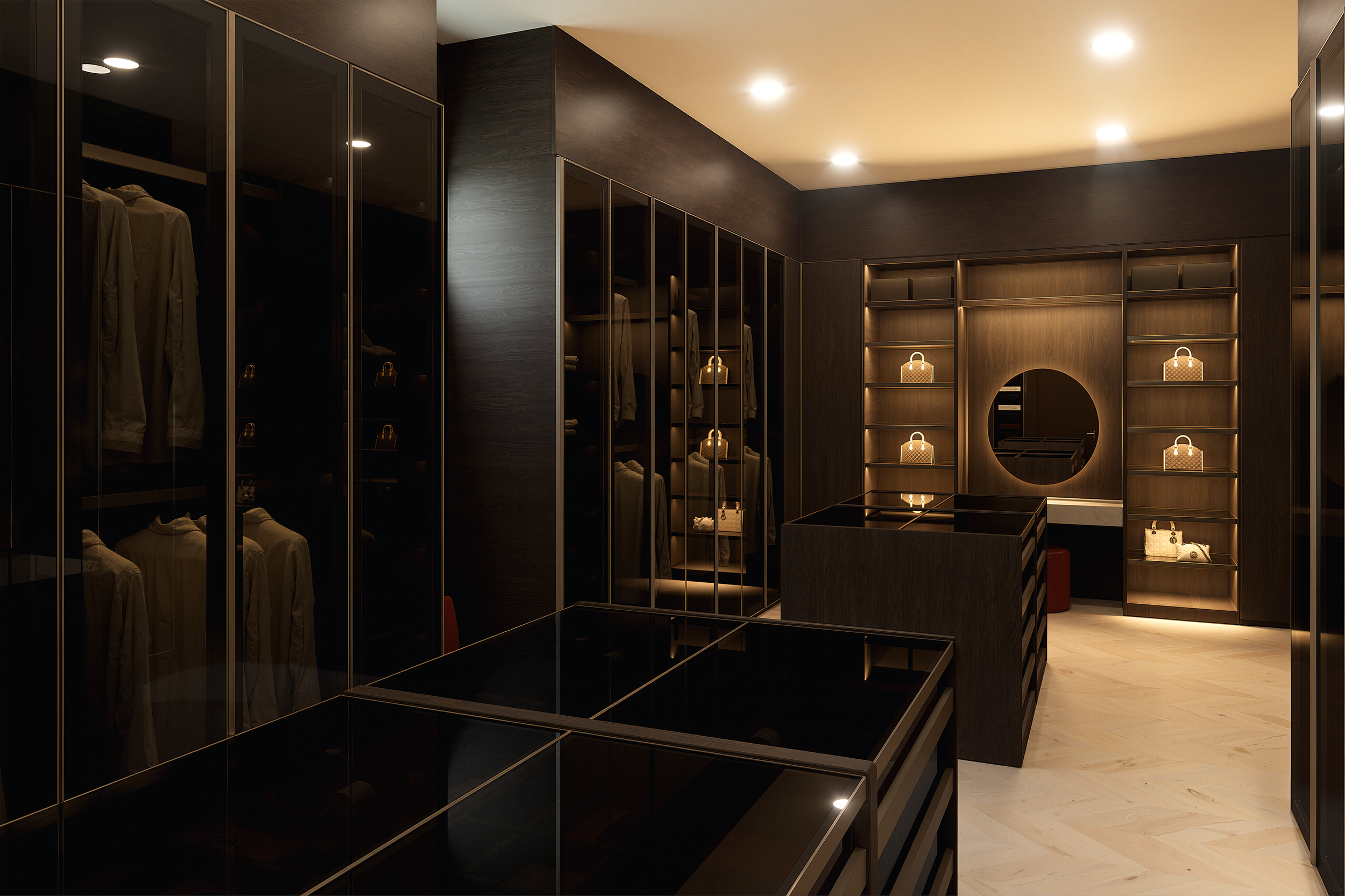 A modern walk-in closet with dark wood and glass cabinets filled with clothes and handbags, illuminated by ceiling lights and wall sconces.