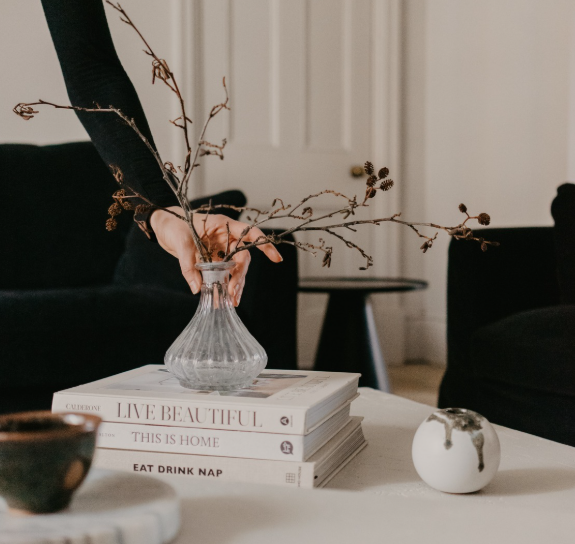 Person arranging dried branches in a glass vase on a stack of books in a cozy living room.