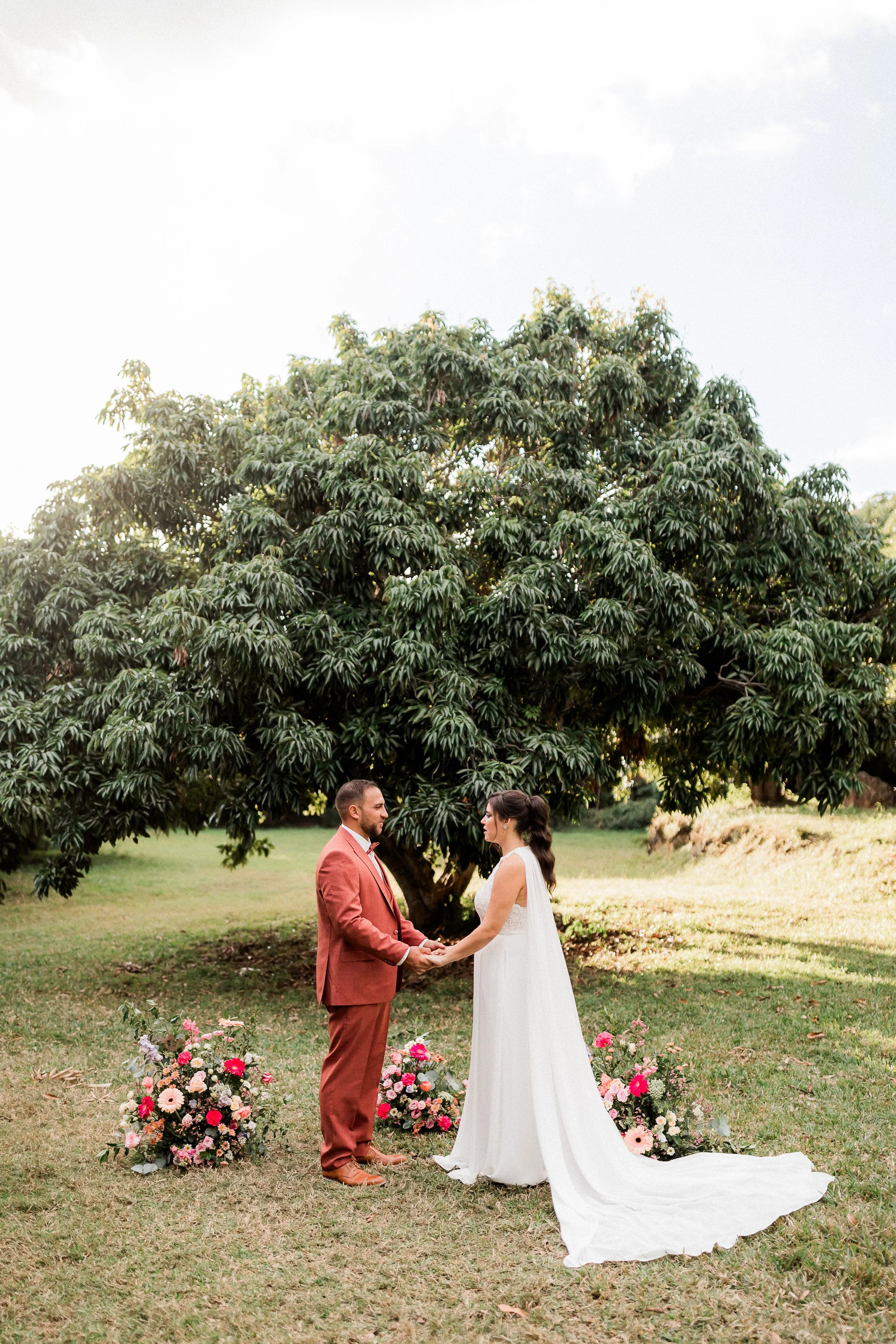 Un couple se tenant la main lors d'une cérémonie de mariage en plein air sous un grand arbre, avec des fleurs colorées disposées au sol.