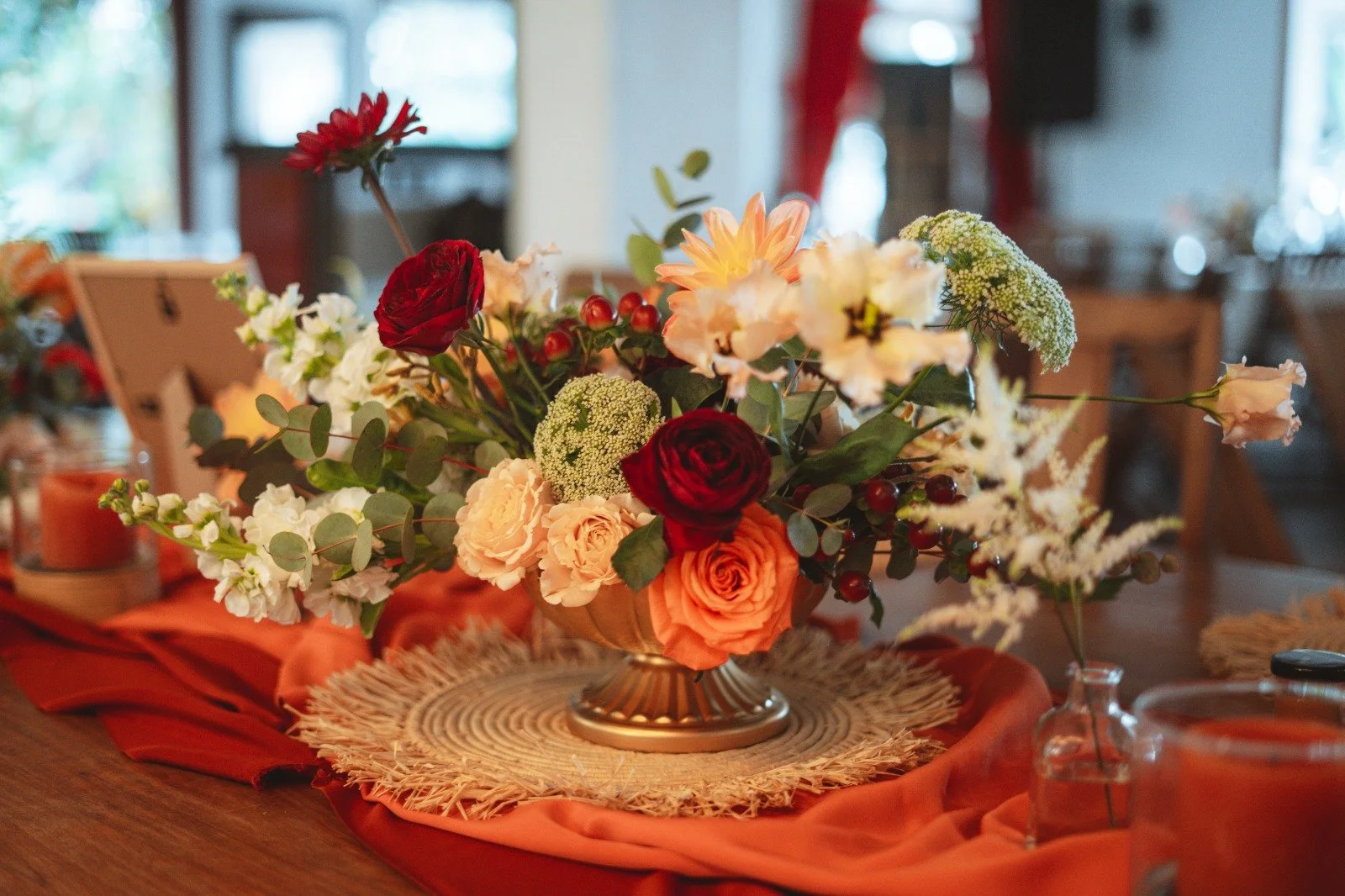 Un arrangement floral avec des roses rouges, orange, roses pâles et autres fleurs, placé dans un vase doré sur une nappe rouge.