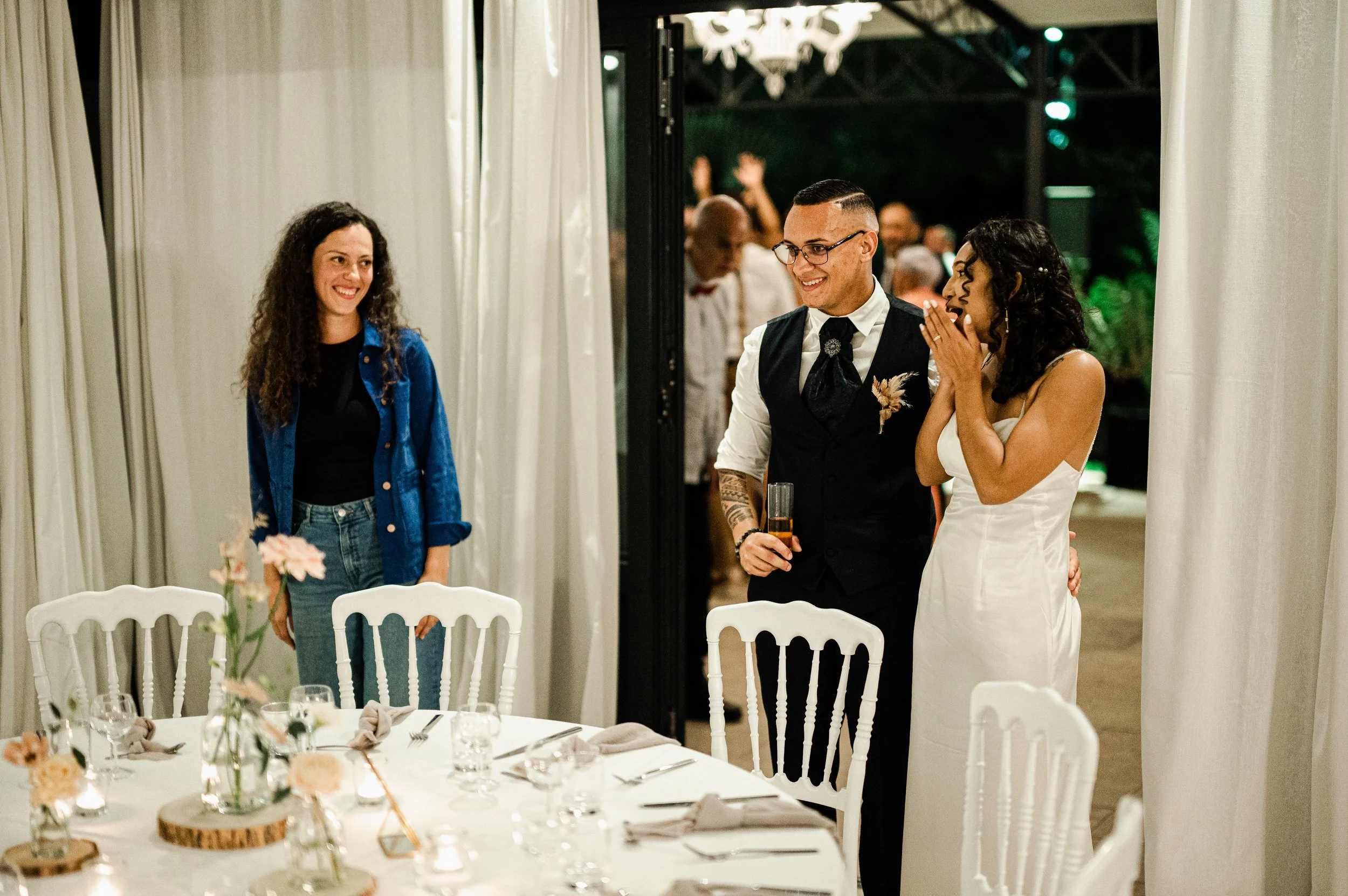 Deux femmes et un homme lors d'une fête ou d'un mariage, avec des invités en arrière-plan, échangeant un moment joyeux.