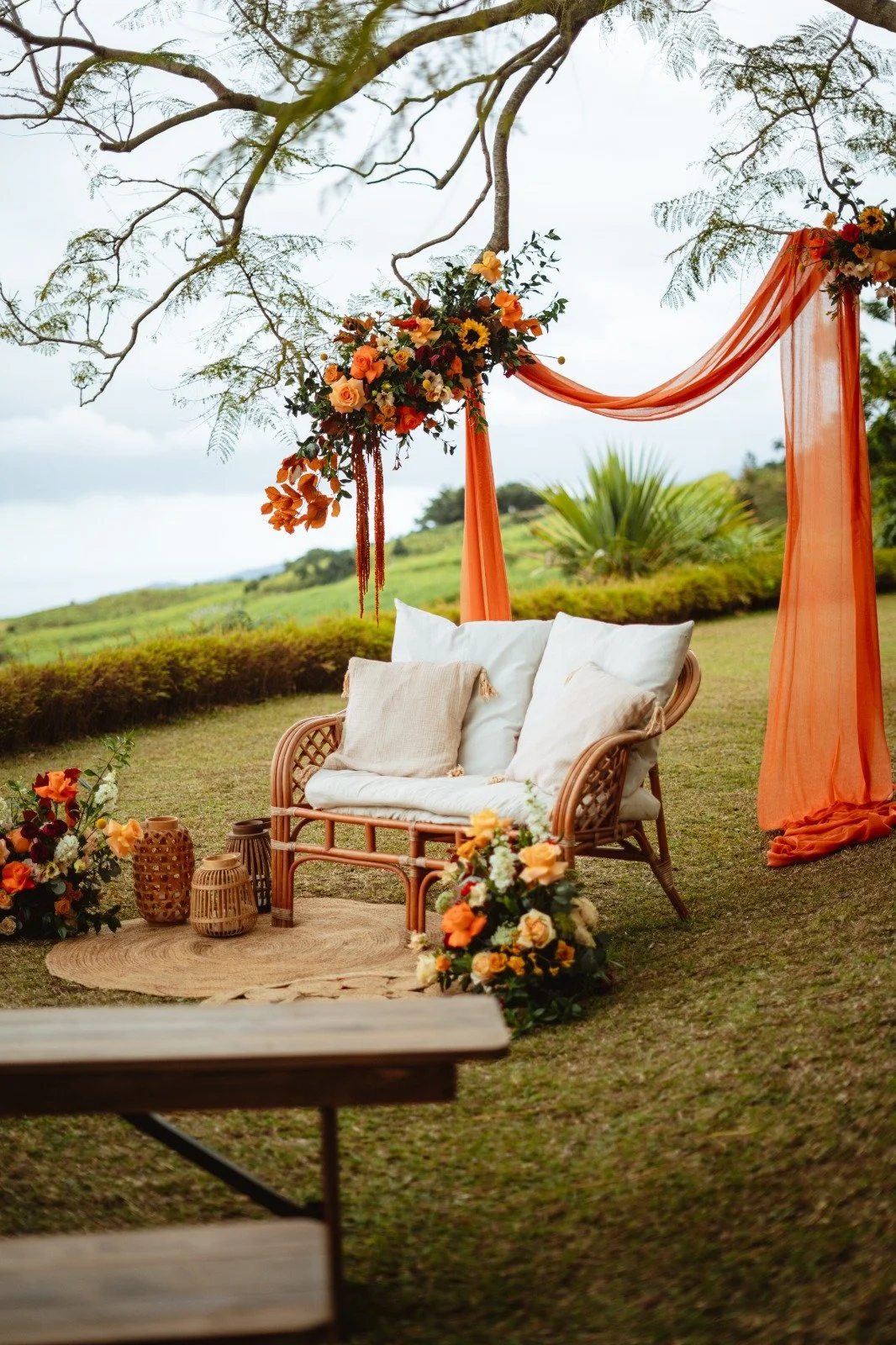 Décoration extérieure avec un canapé en rotin, coussins beiges, fleurs oranges et blanches, draperies orange, dans un espace naturel avec arbre.