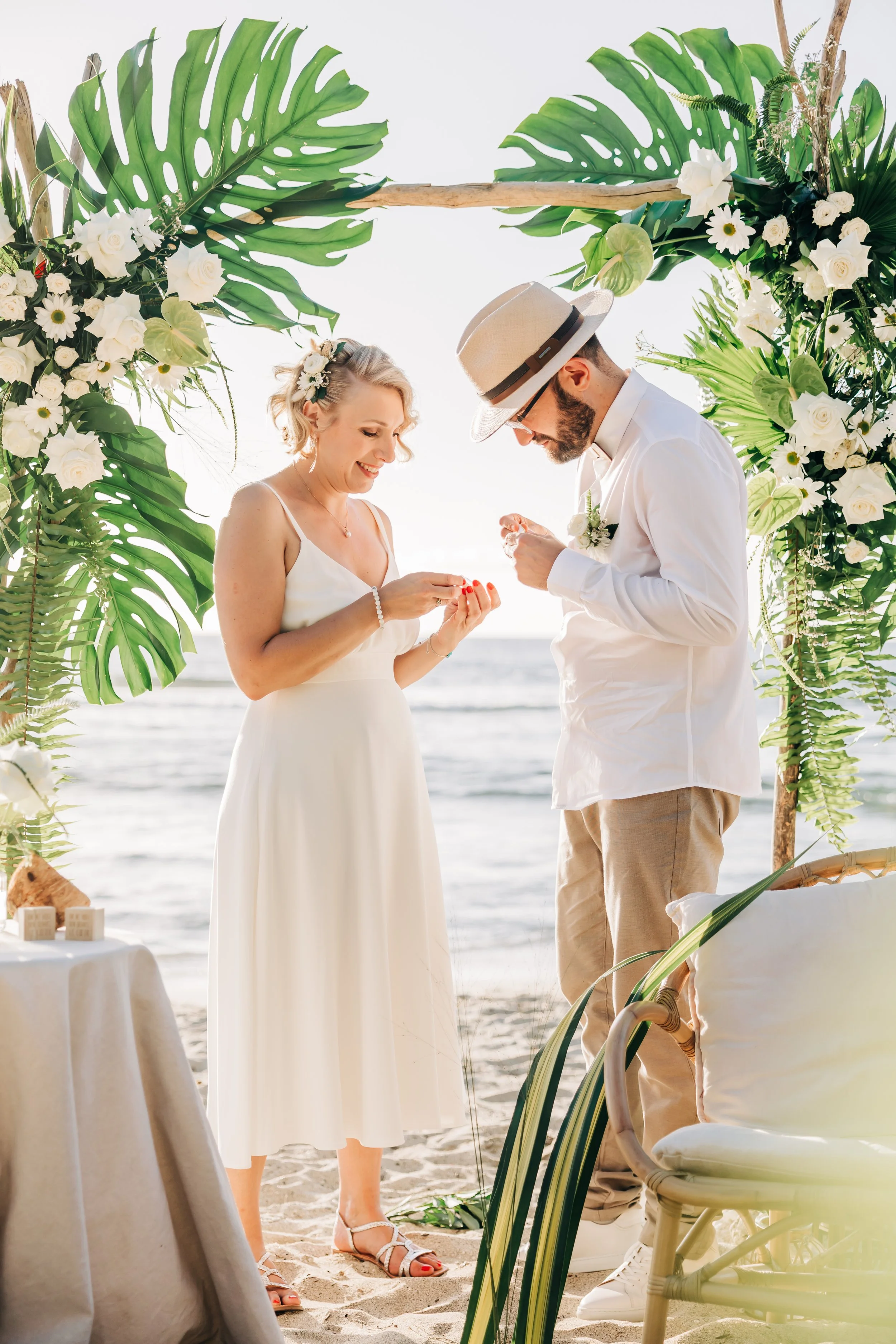 Un couple échange des vœux lors d'une cérémonie de mariage sur la plage, entouré de fleurs blanches et de feuilles vertes, avec la mer en arrière-plan.
