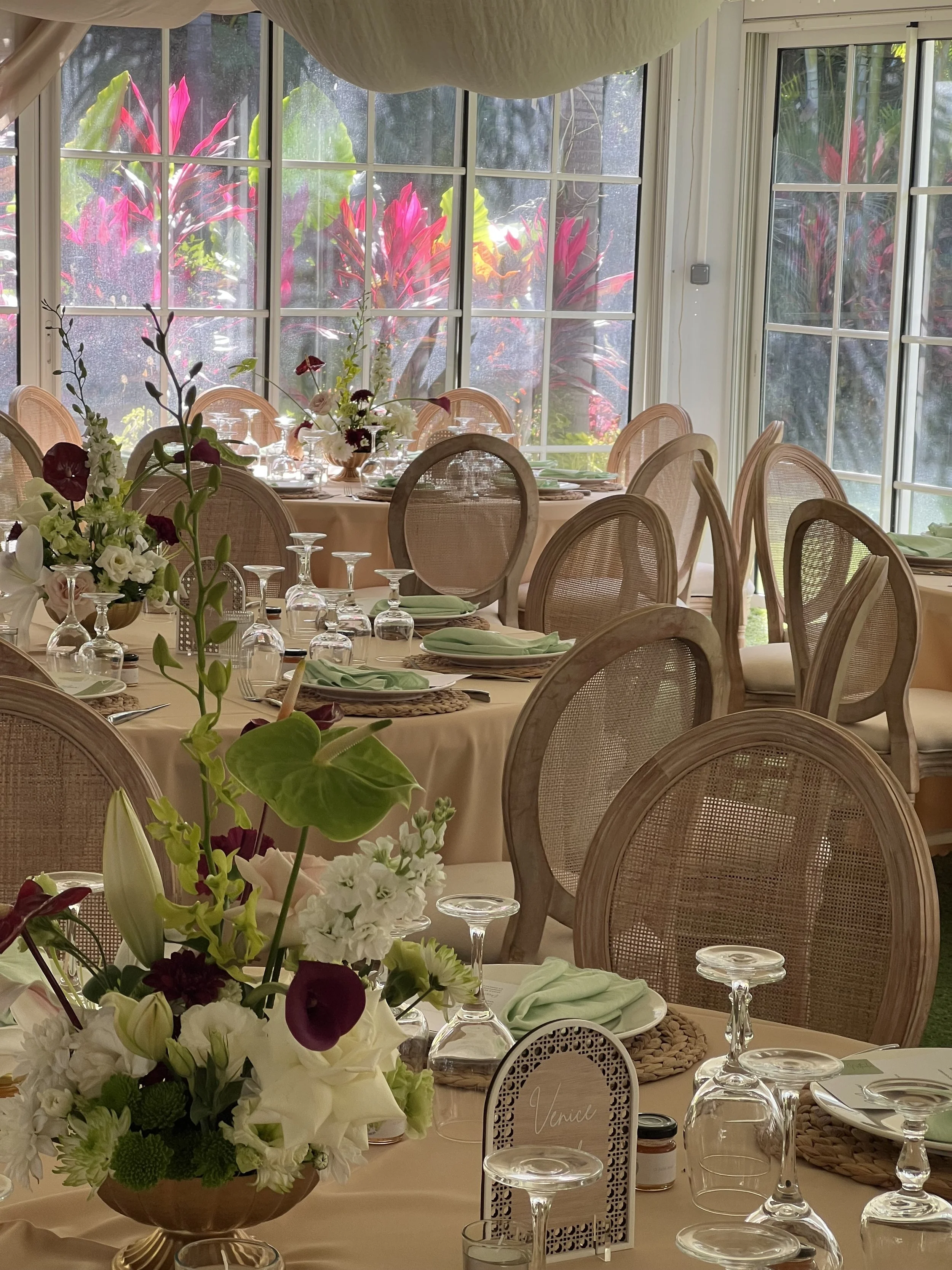 Table décorée avec des fleurs blanches, roses et violettes, prête pour un repas. Fenêtres grandes laissant entrer la lumière naturelle, en arrière-plan des plantes tropicales.