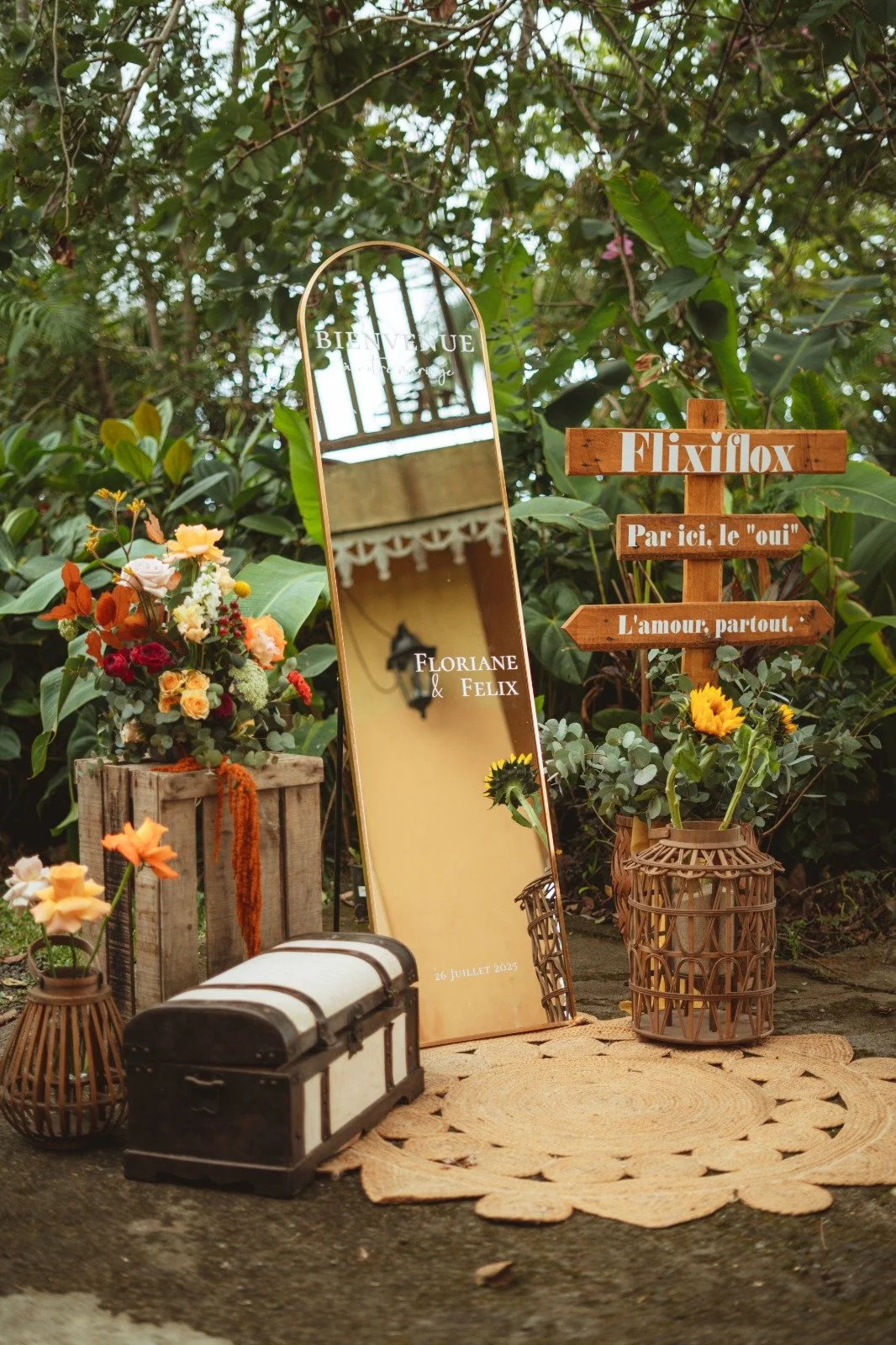 Décoration de mariage en extérieur avec un miroir, des fleurs, un coffre en bois et un panneau en bois avec des inscriptions, ambiance champêtre.