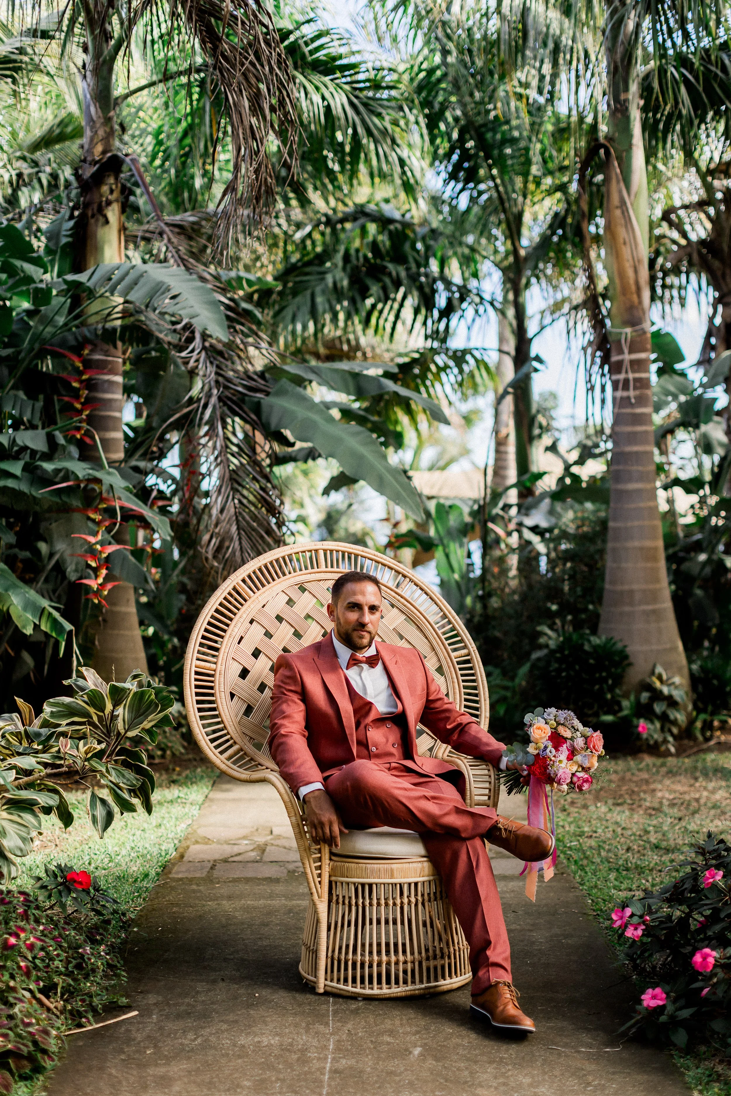 Un homme en costume marron assis dans une chaise en rotin dans un jardin luxuriant avec des fleurs colorées.