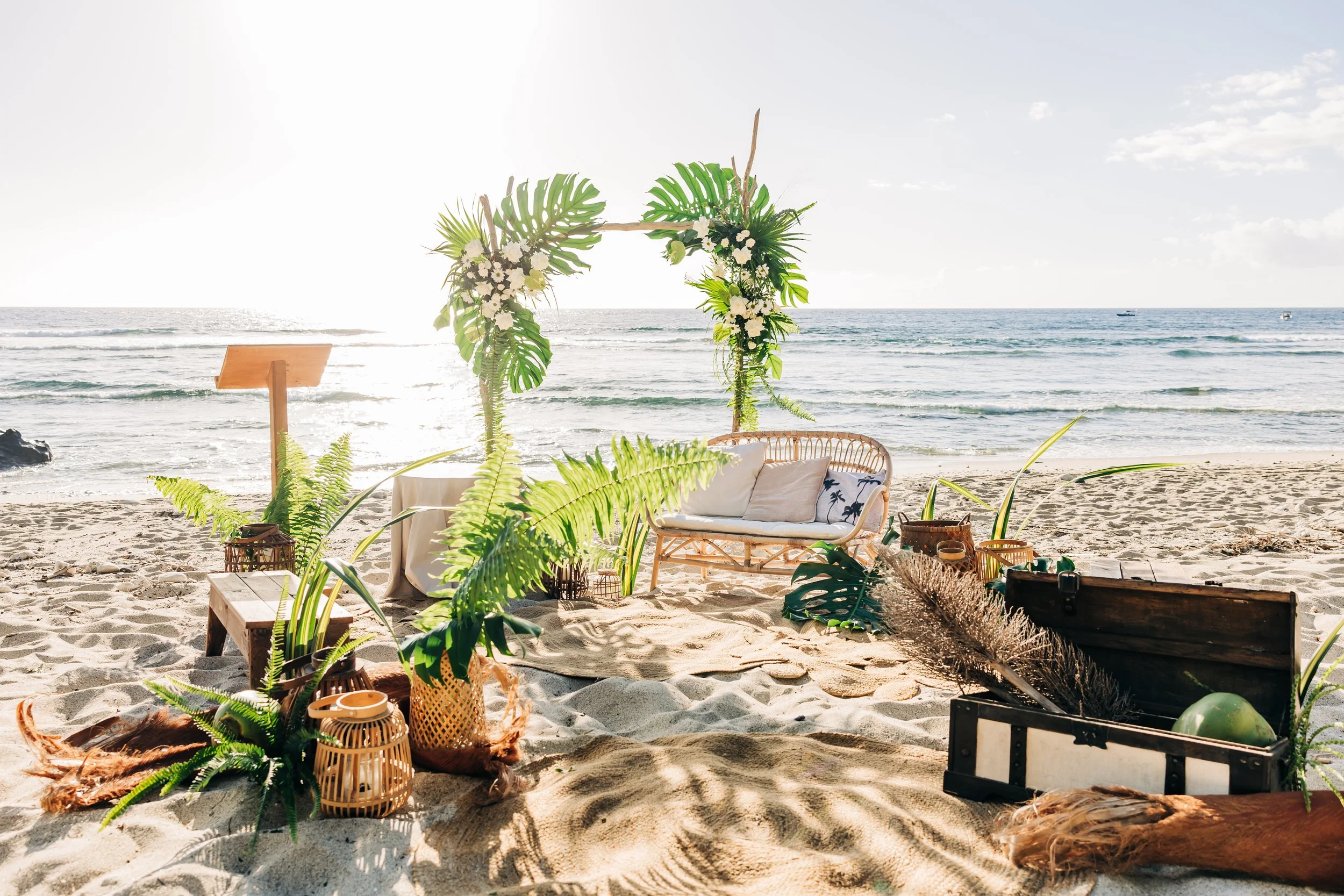 Décor de mariage sur la plage avec une arche en fleurs et feuilles vertes, mobilier en rotin, et vue sur la mer et le ciel ensoleillé.
