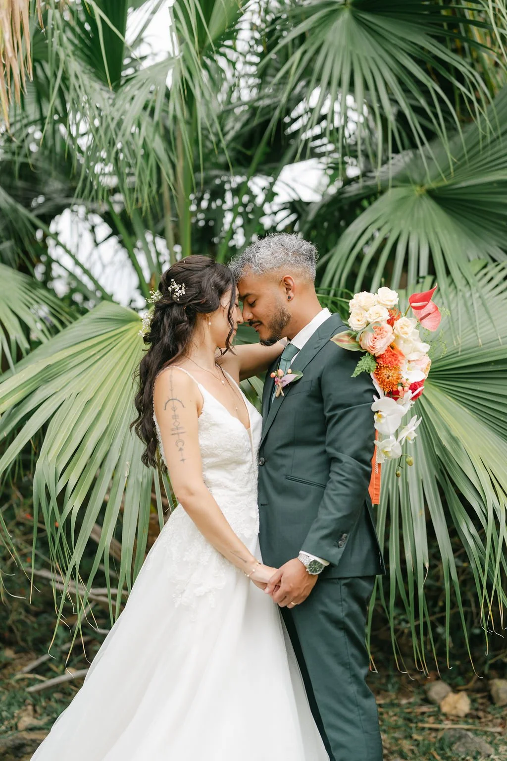 Un couple marié partage un moment intime dans un environnement de plantes vertes, la femme porte une robe de mariage blanche, et l'homme un costume sombre. La femme tient un bouquet de fleurs colorées.