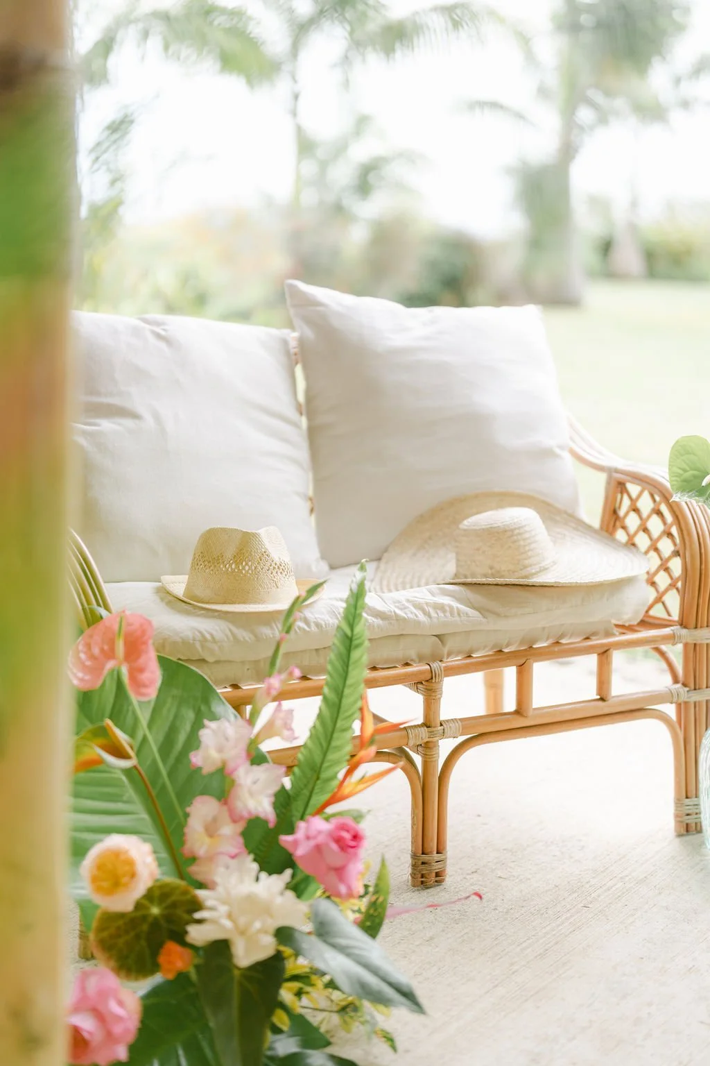 Un canapé en rotin avec des coussins blancs, un chapeau de paille posé dessus, et des fleurs colorées au premier plan dans un jardin extérieur ensoleillé.