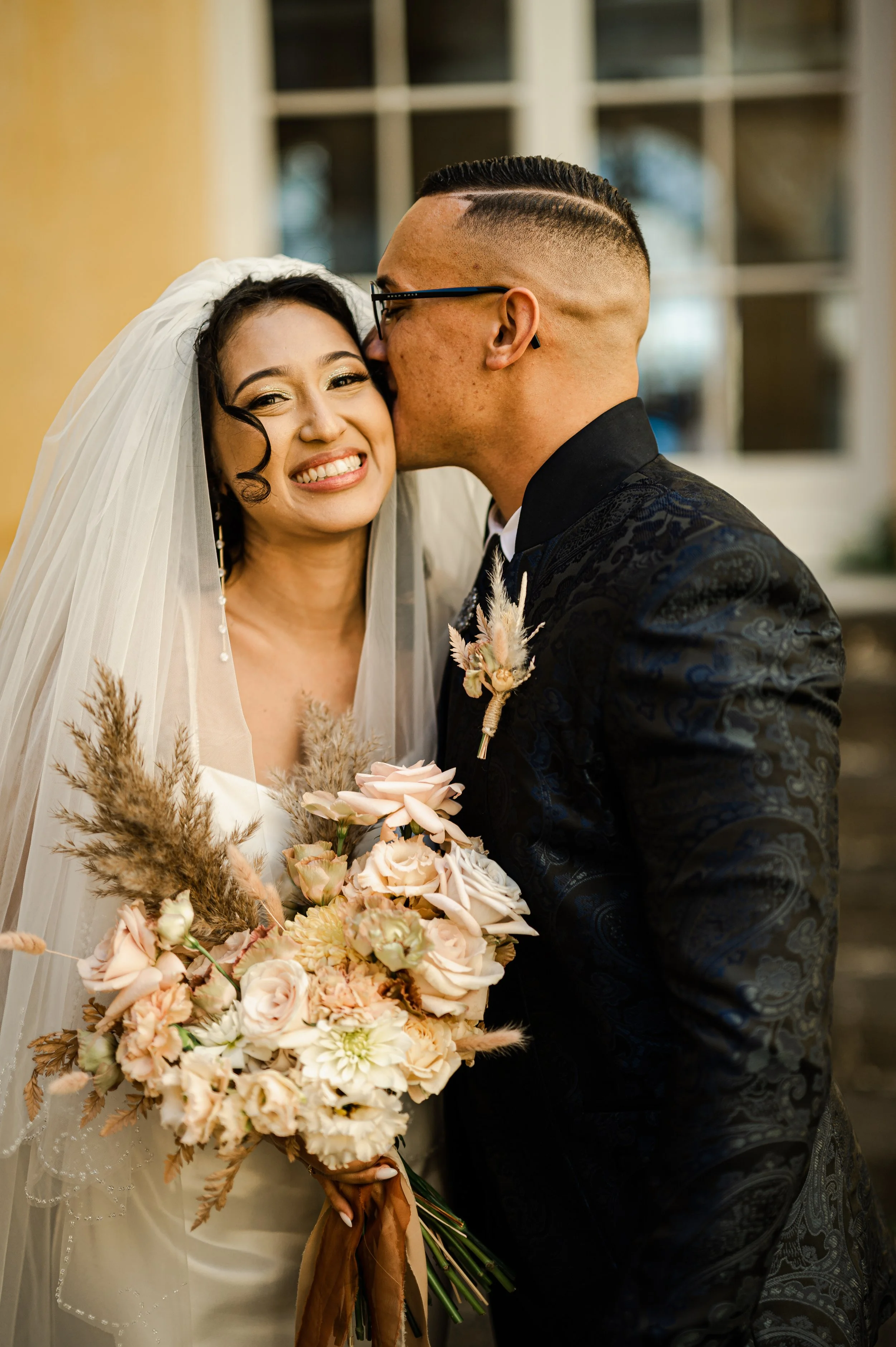 Un couple de mariés lors d'une cérémonie de mariage, la femme souriante porte une robe blanche avec un voile, et tient un bouquet de fleurs, pendant que l'homme l'embrasse sur la joue, portant une veste noire avec des broderies et des lunettes.